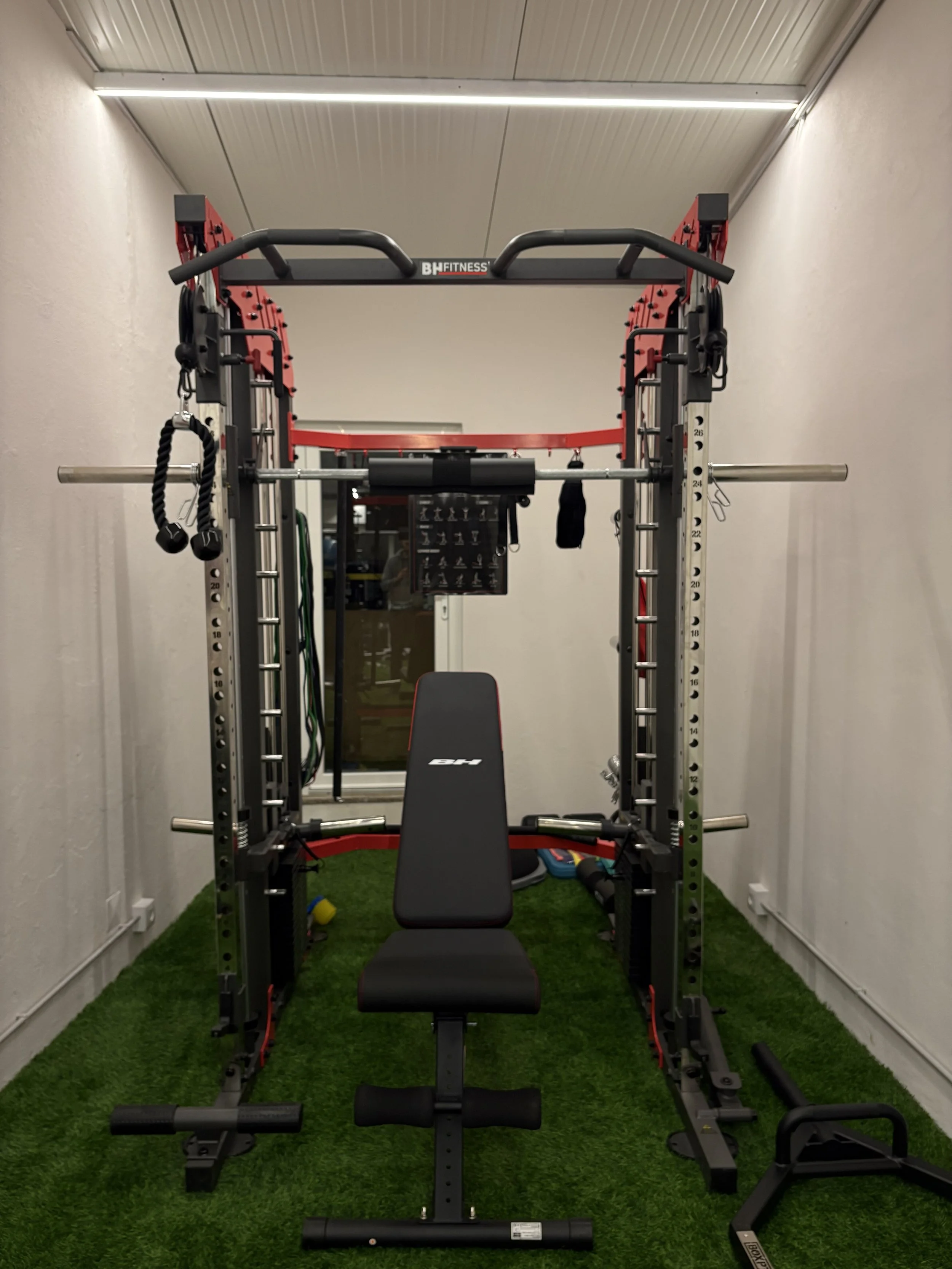 Estrutura de academia com barra para levantamento de peso, banco de exercícios e equipamentos de treino em um espaço com piso de grama artificial.