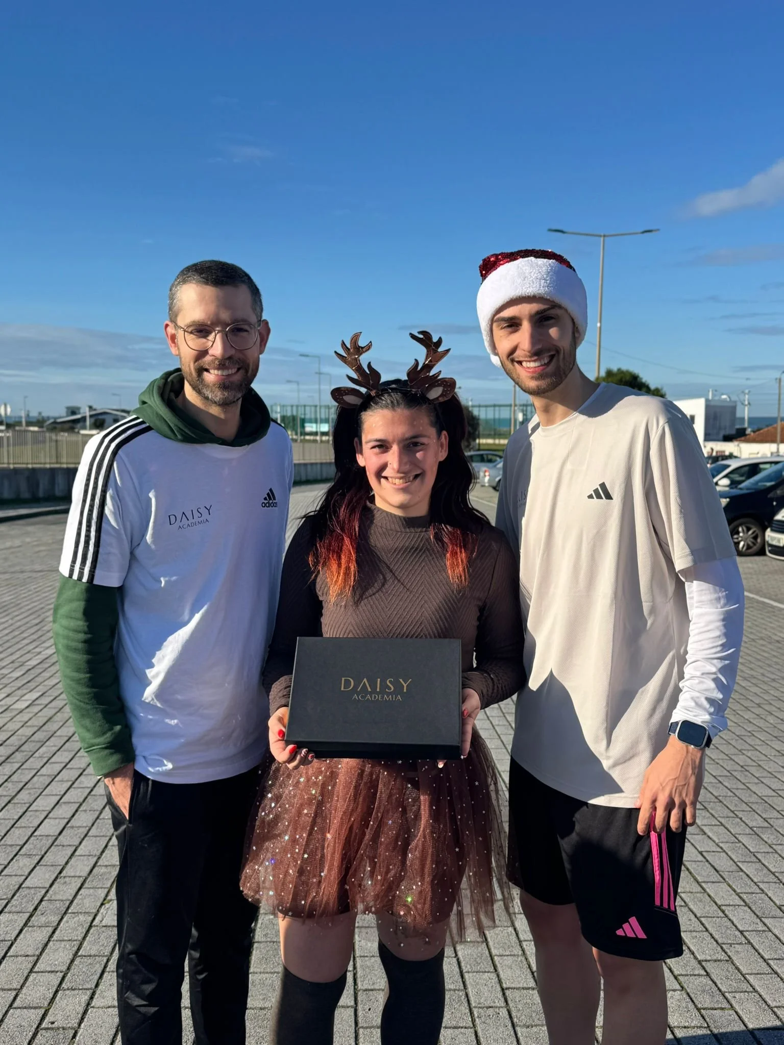 Grupo de três pessoas sorrindo ao ar livre, celebrando, com uma mulher no centro segurando uma caixa da Daisy Academia. Ela usa uma fantasia de Natal com chifres de rena, enquanto um homem do lado direito usa um gorro de Papai Noel, e o outro homem d