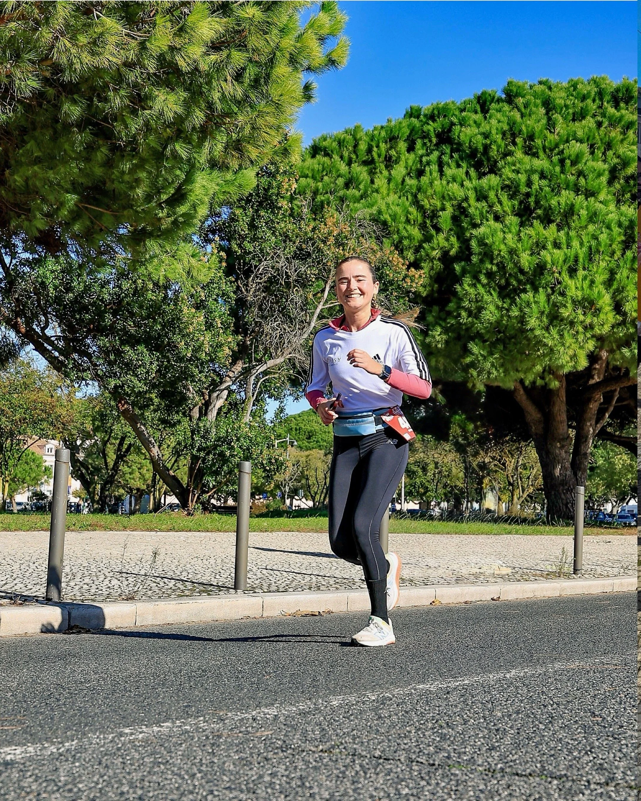 Mulher correndo na rua, usando roupas esportivas, sorrindo, com árvores verdes ao fundo e céu azul.