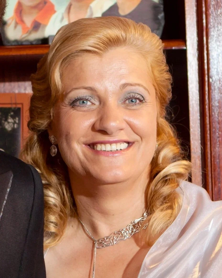 Mulher com cabelo loiro claro, vestindo maquiagem leve, usando brinco e colar prateado, sorrindo para a câmera, com fundo de uma estante com fotos.