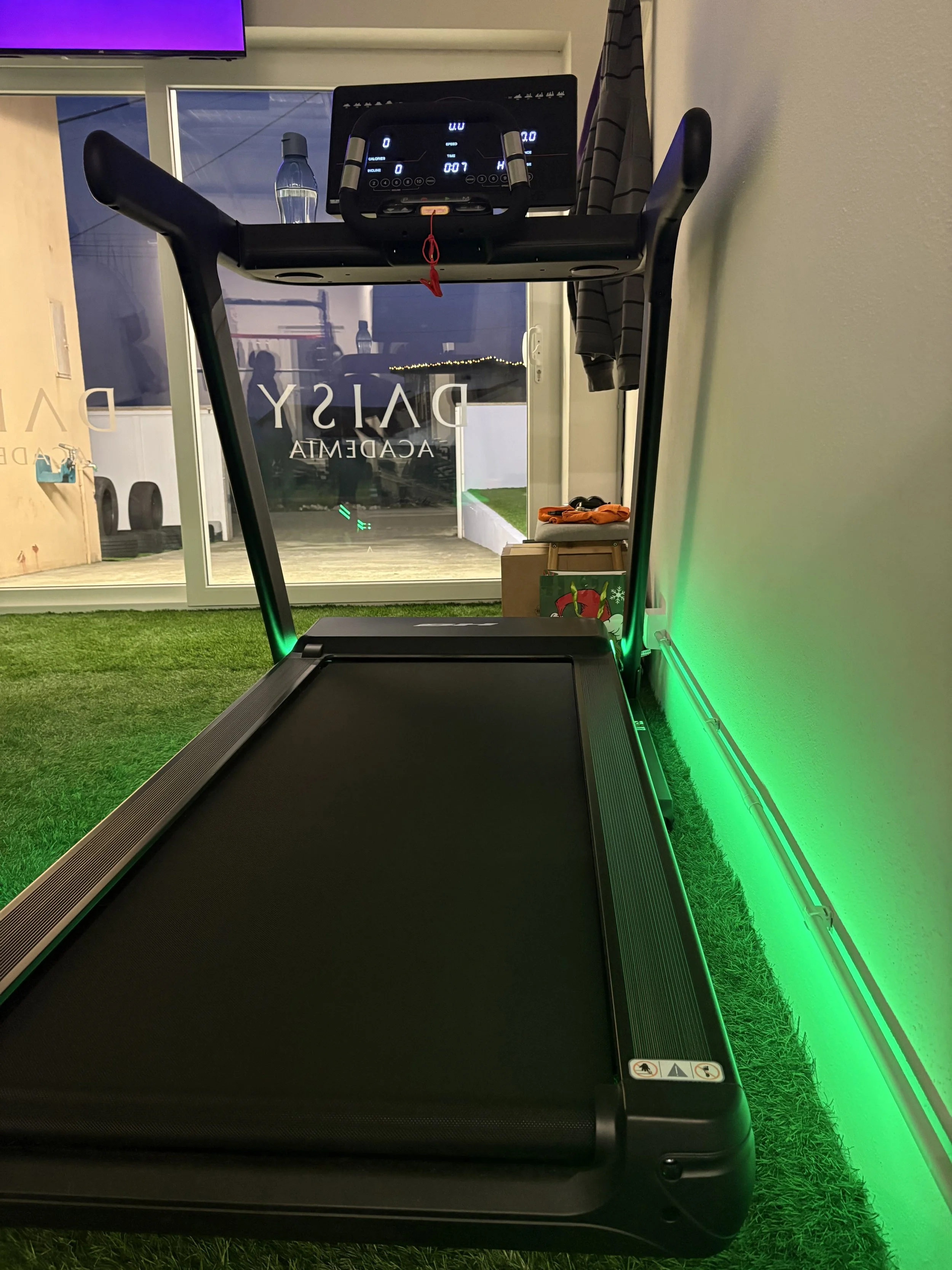 Este é um aparelho de esteira de corrida em um ambiente interno com piso de grama artificial, com iluminação verde na parede ao lado. Ao fundo, uma porta de vidro revela uma área ao ar livre.