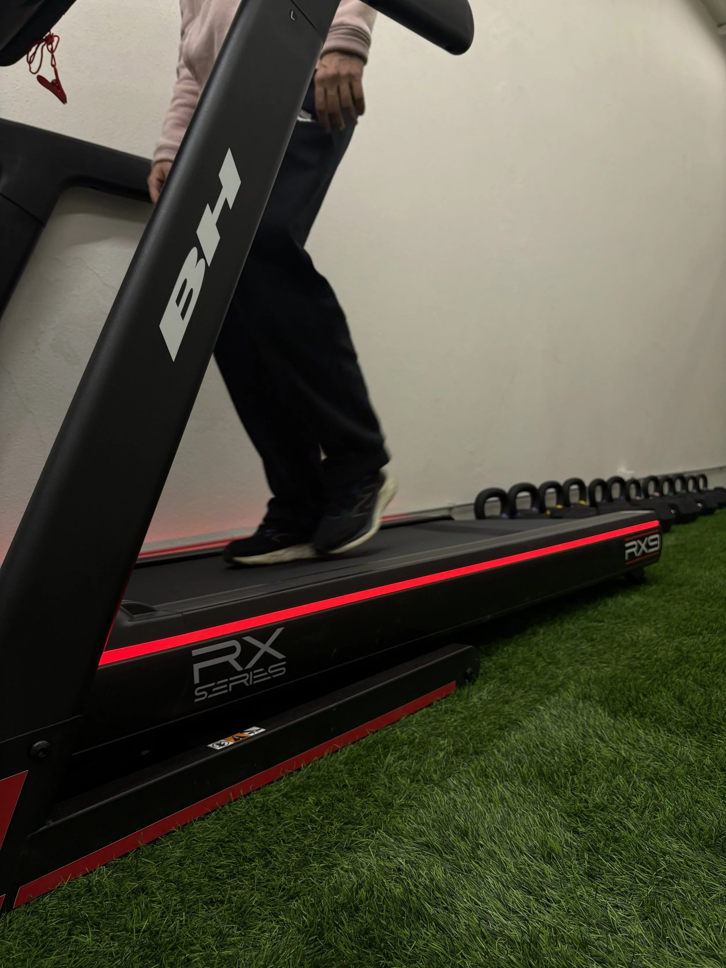 Pessoa caminhando na esteira elétrica RX Series, localizada sobre uma área de grama artificial, com alguns kettlebells alinhados no fundo.