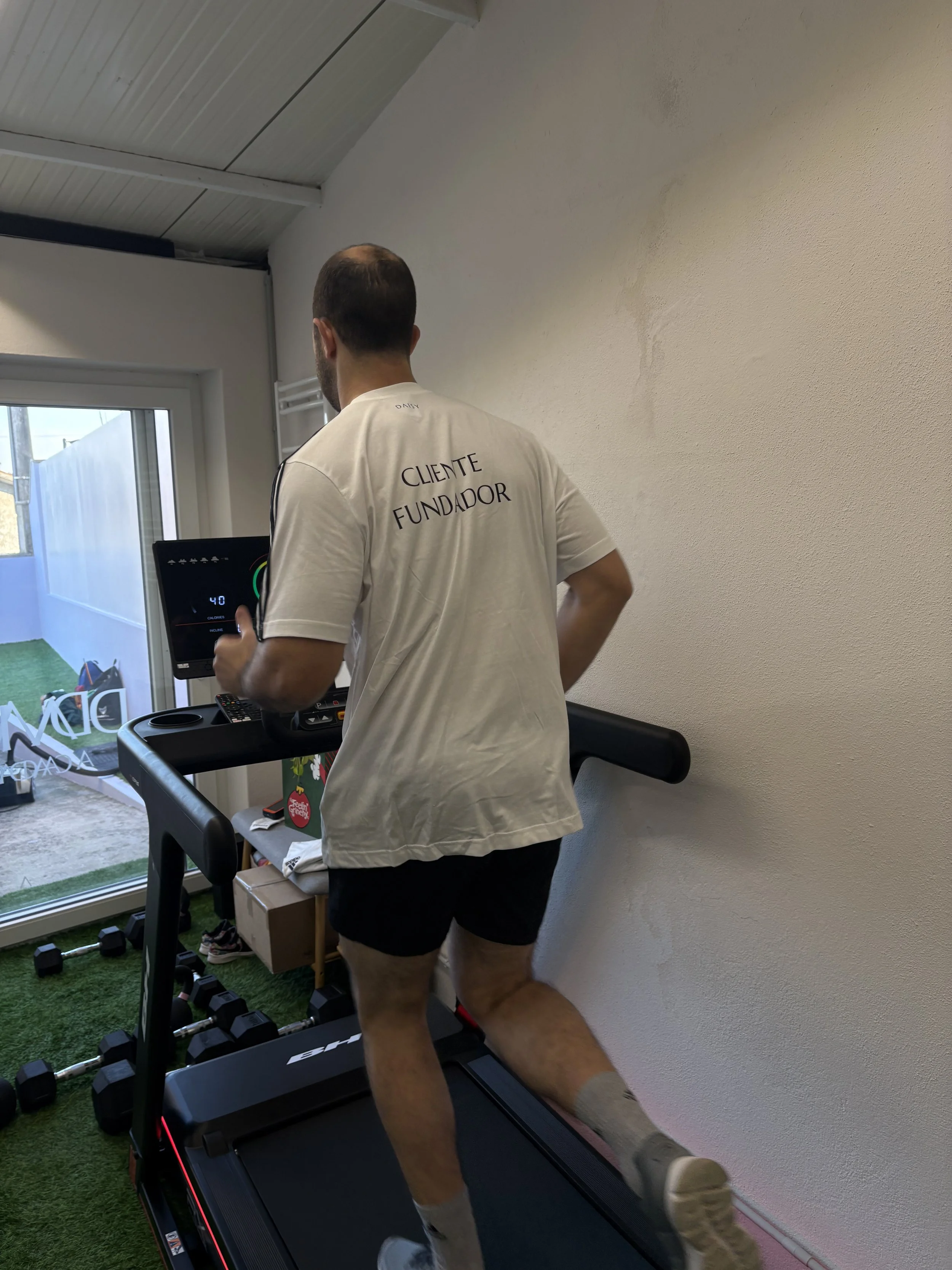 Homem correndo na esteira em uma academia, usando camiseta com a frase 'Cliente Fundador' nas costas e shorts pretos.