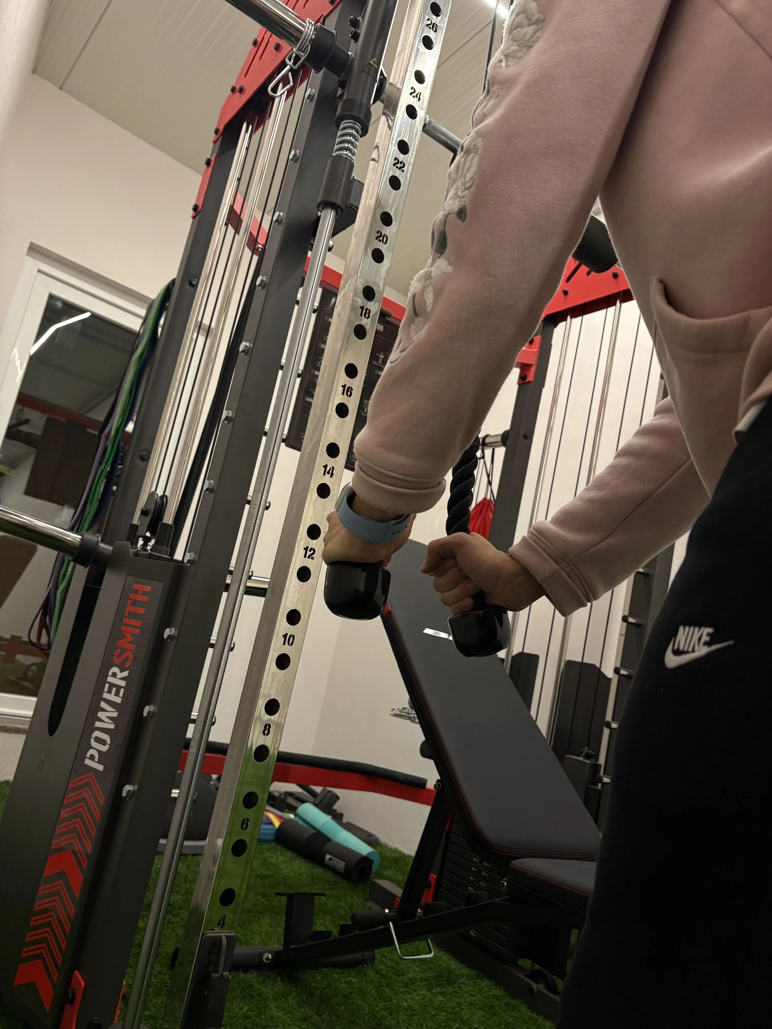 Pessoa segurando uma corda de puxar em uma academia, com peso de 20 kg, ao lado de um aparelho de treino vermelho e preto, protocolo Power Smith, em um ambiente de academia com piso de grama artificial.