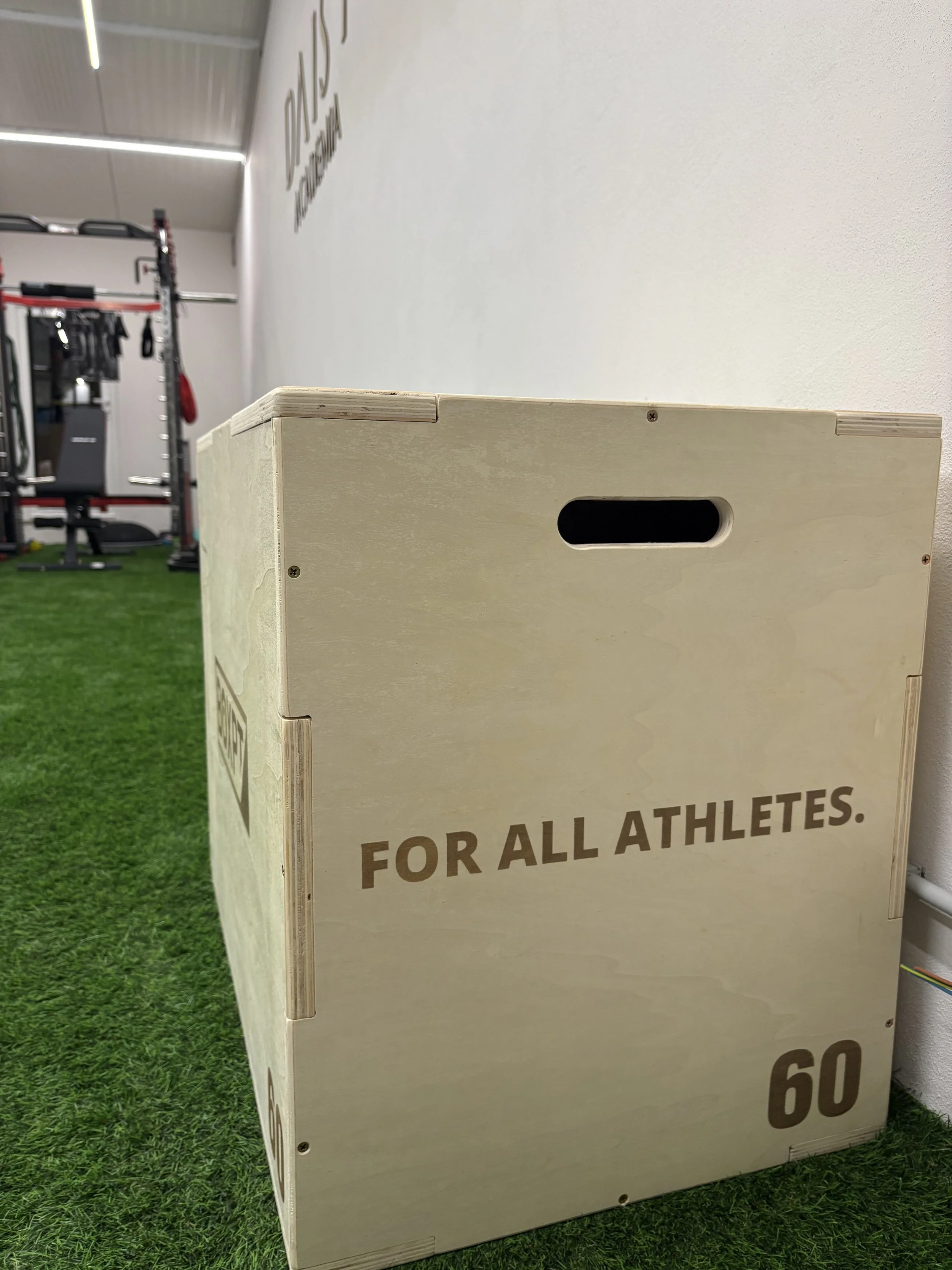 Caixa de treino de madeira no ginásio com a inscrição 'For All Athletes' e o número 60, ao lado de grama artificial e equipamentos de ginástica ao fundo.