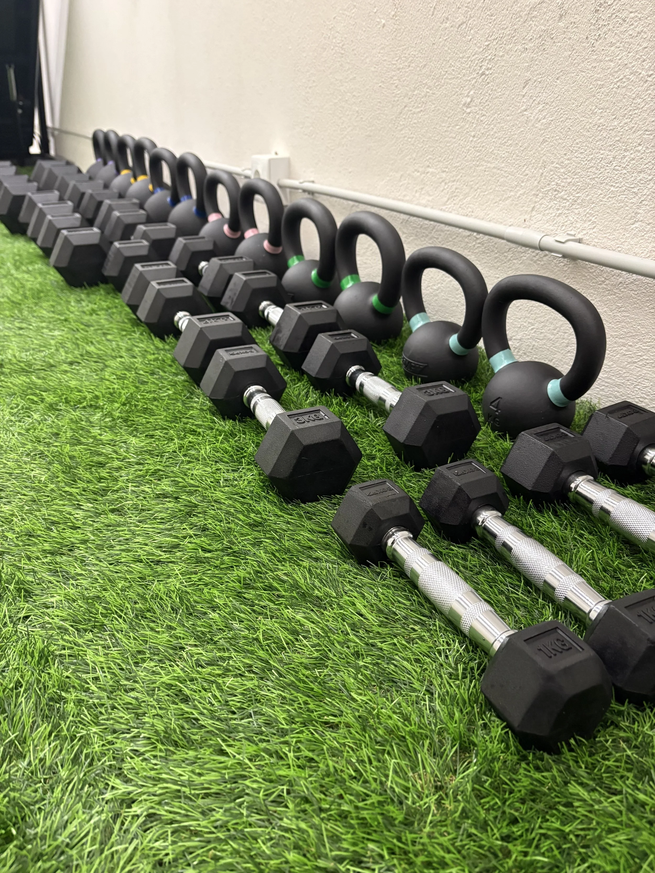 Conjunto de halteres e kettlebells alinhados no chão de grama sintética em uma academia.