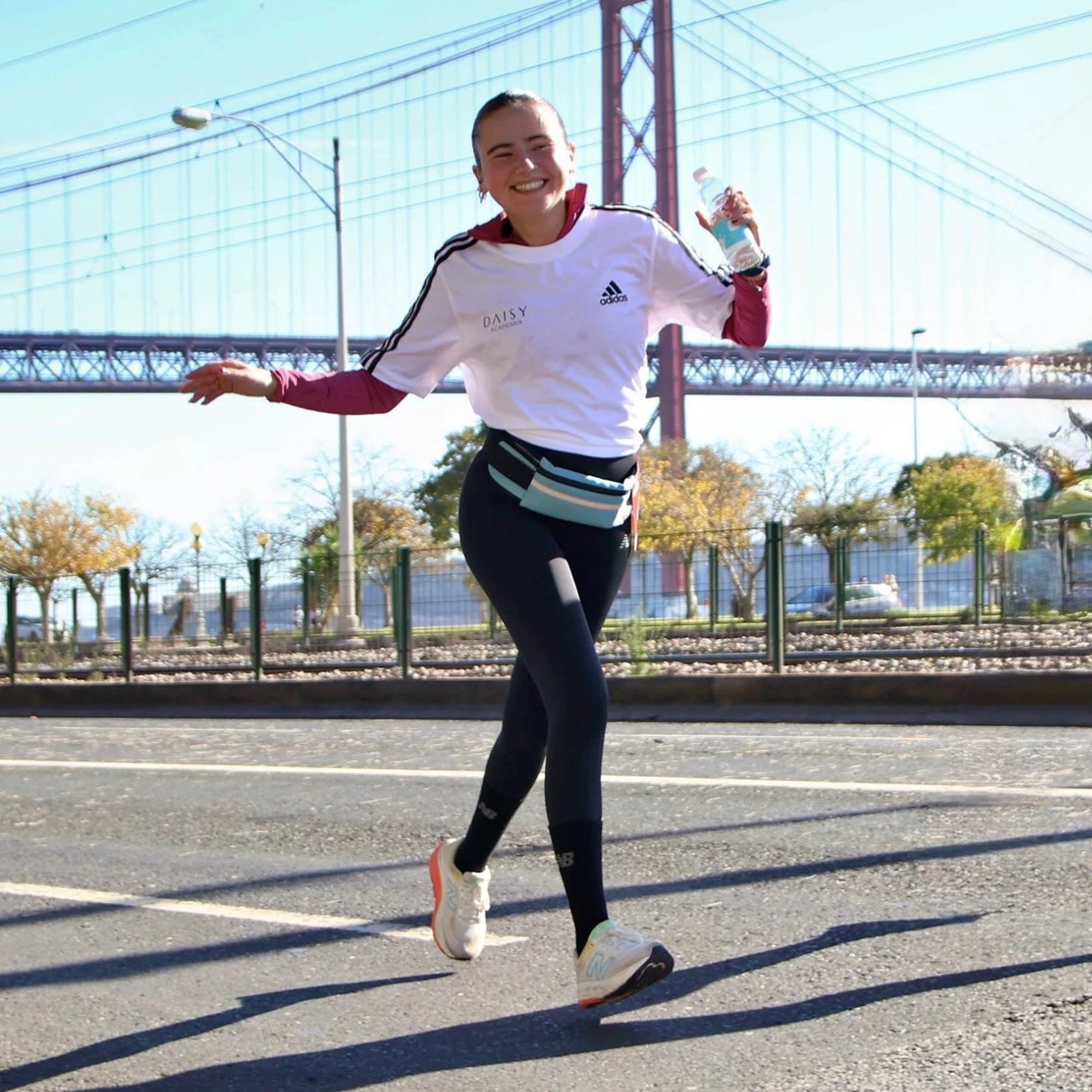 Jovem sorridente fazendo corrida na rua com ponte ao fundo, vestida com roupas esportivas, usando um top com jaqueta branca por baixo, calças pretas, tênis branco, e carregando uma garrafa de água.