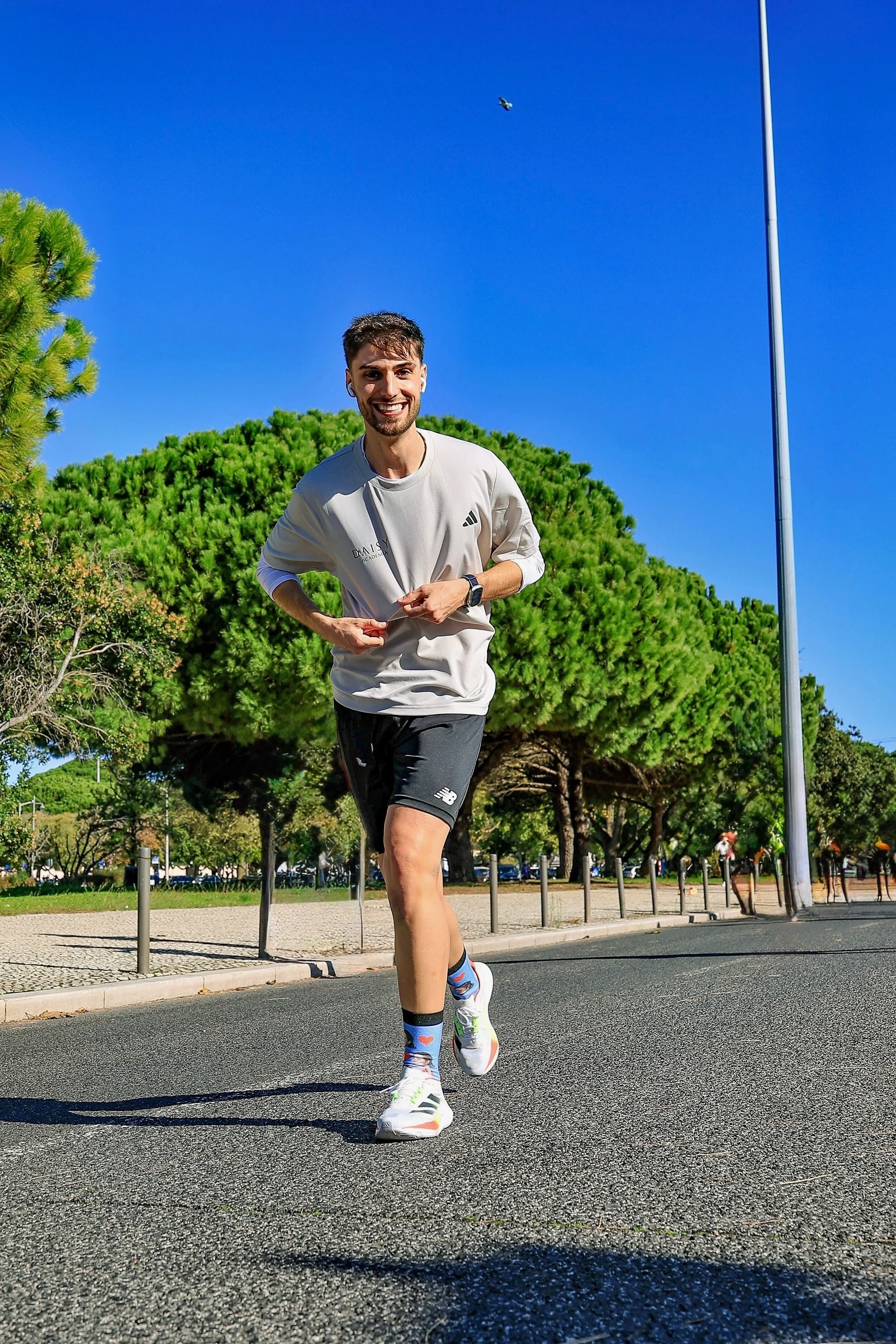 Homem correndo na rua durante o dia, com árvores verdes, céu azul claro e poste de luz ao fundo.