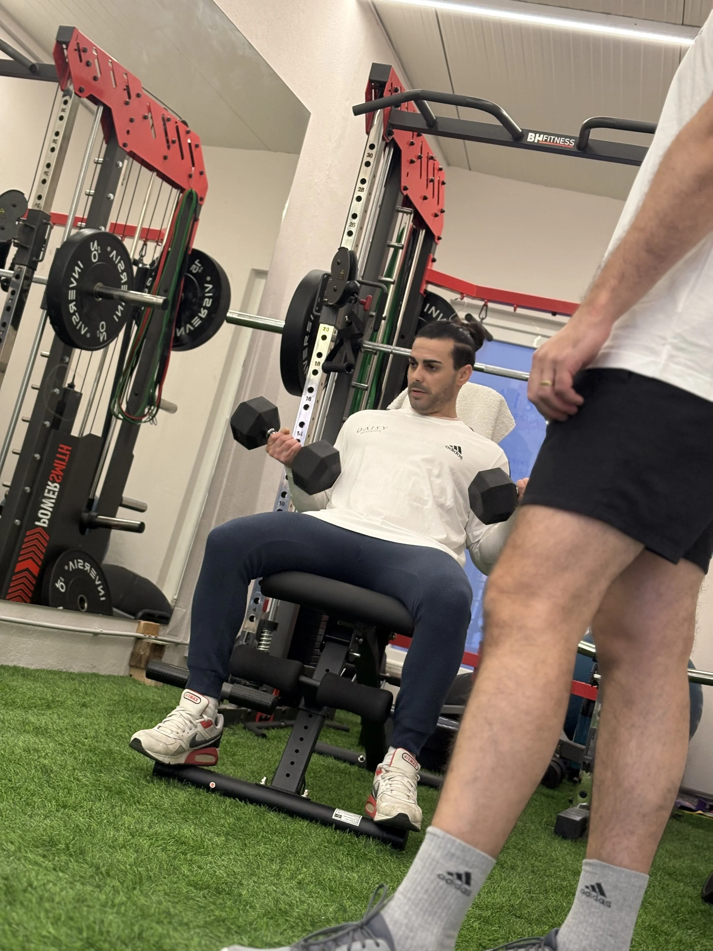 Pessoa fazendo exercício de bíceps com halteres enquanto está sentado em um banco na academia, com um treinador ao lado observando.