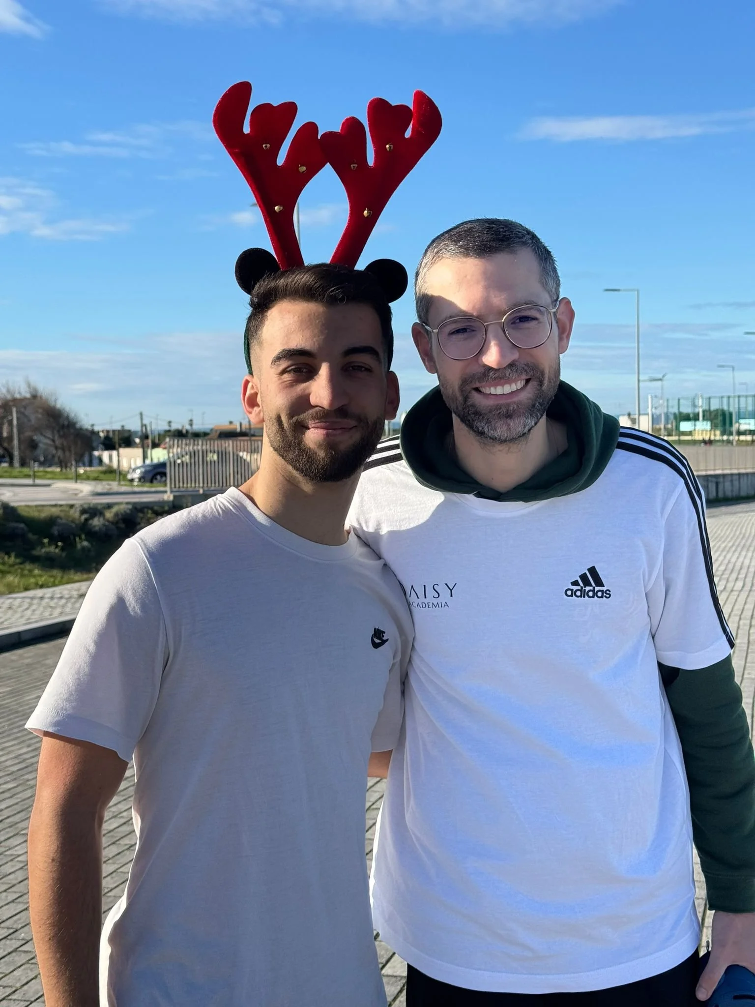 Homens sorrindo ao ar livre, um de cabelos escuros e barba, o outro de cabelos cinzentos e barba, usando óculos e camisetas brancas, um deles com adereço de rena na cabeça