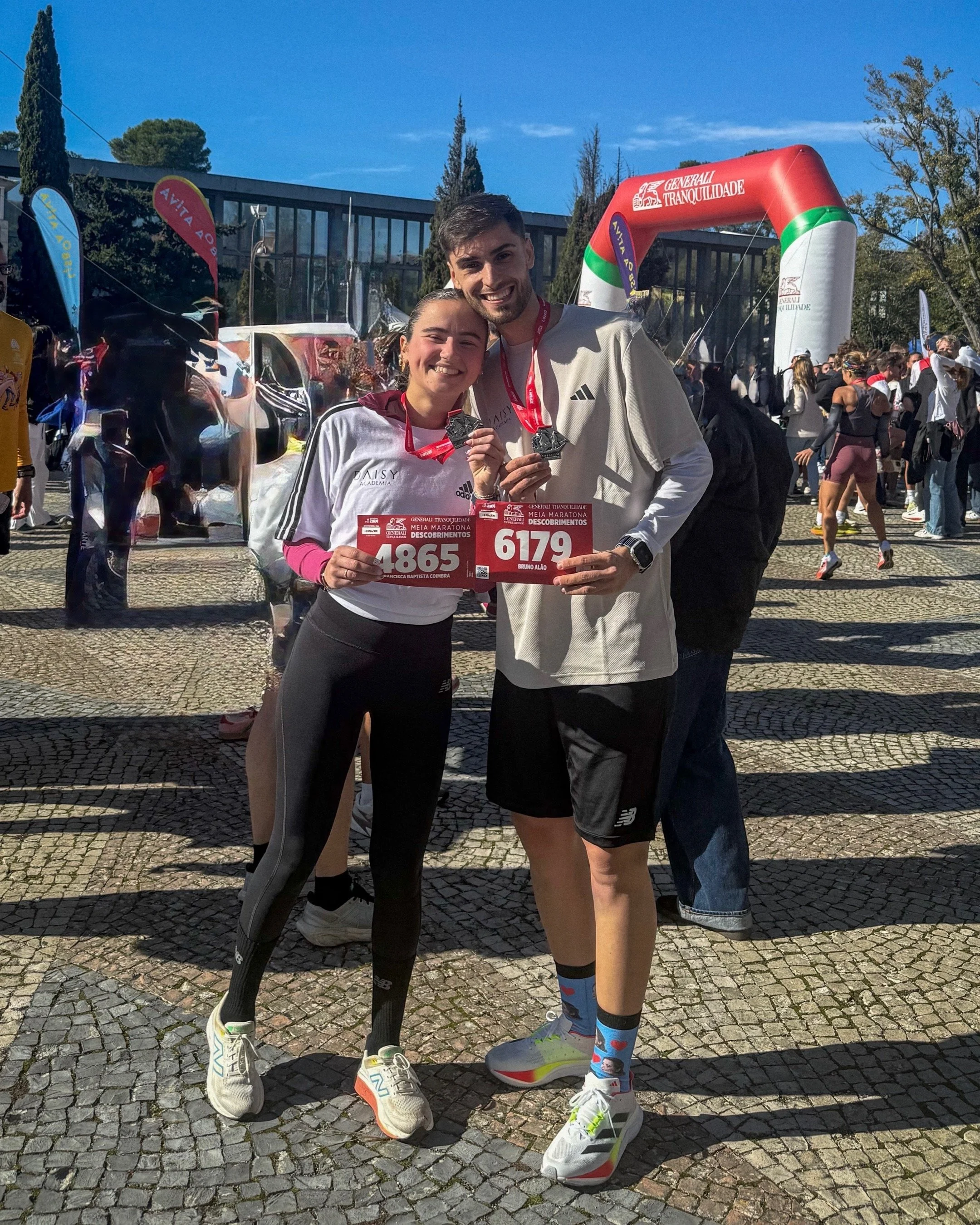 Dois corredores sorridentes segurando medalhas e placas de participação após uma corrida em um evento ao ar livre, com uma arquibancada inflável ao fundo e muitas pessoas ao redor.