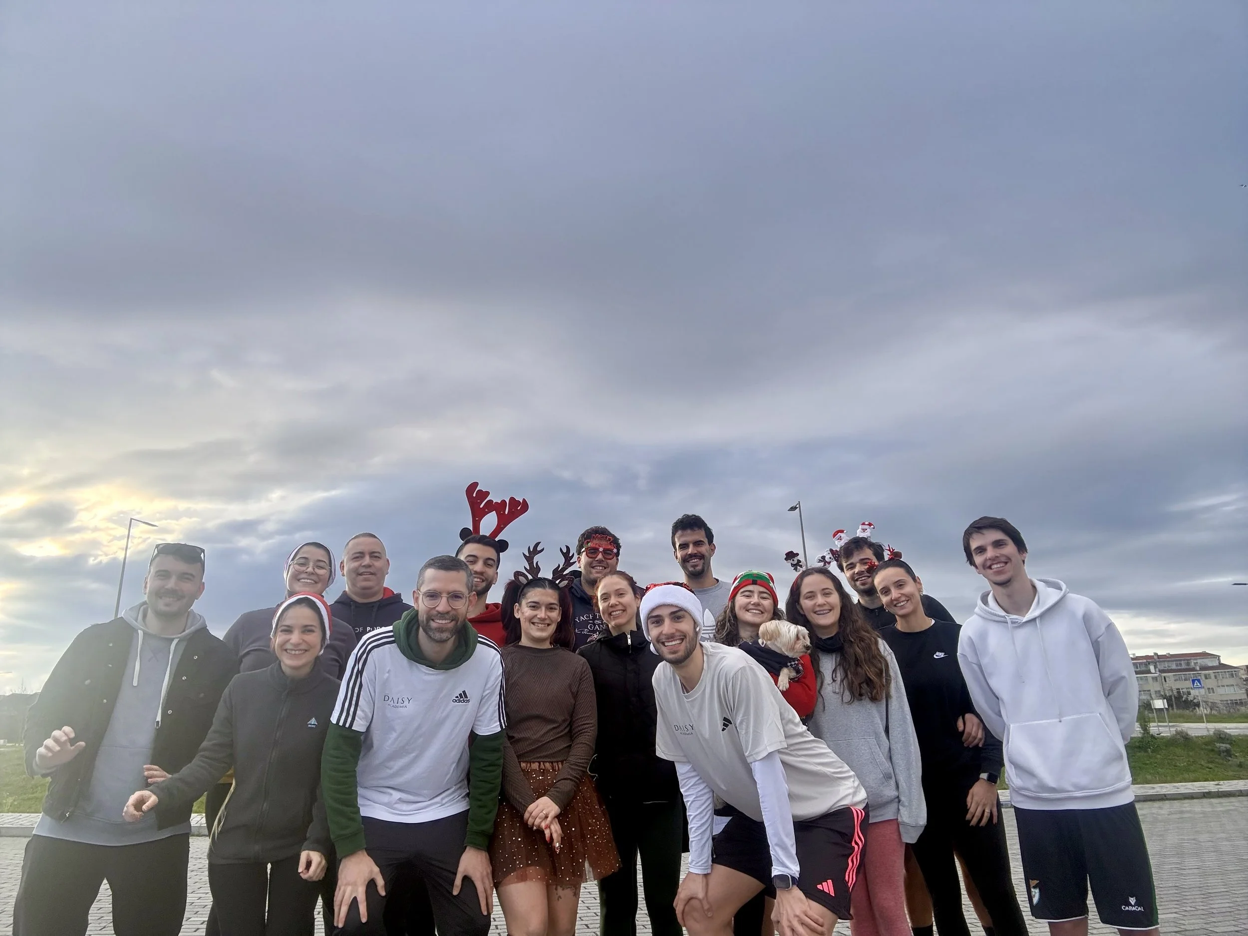 Grupo de pessoas sorrindo ao ar livre, alguns usando acessórios natalinos, como gorros de Papai Noel e chifres de rena