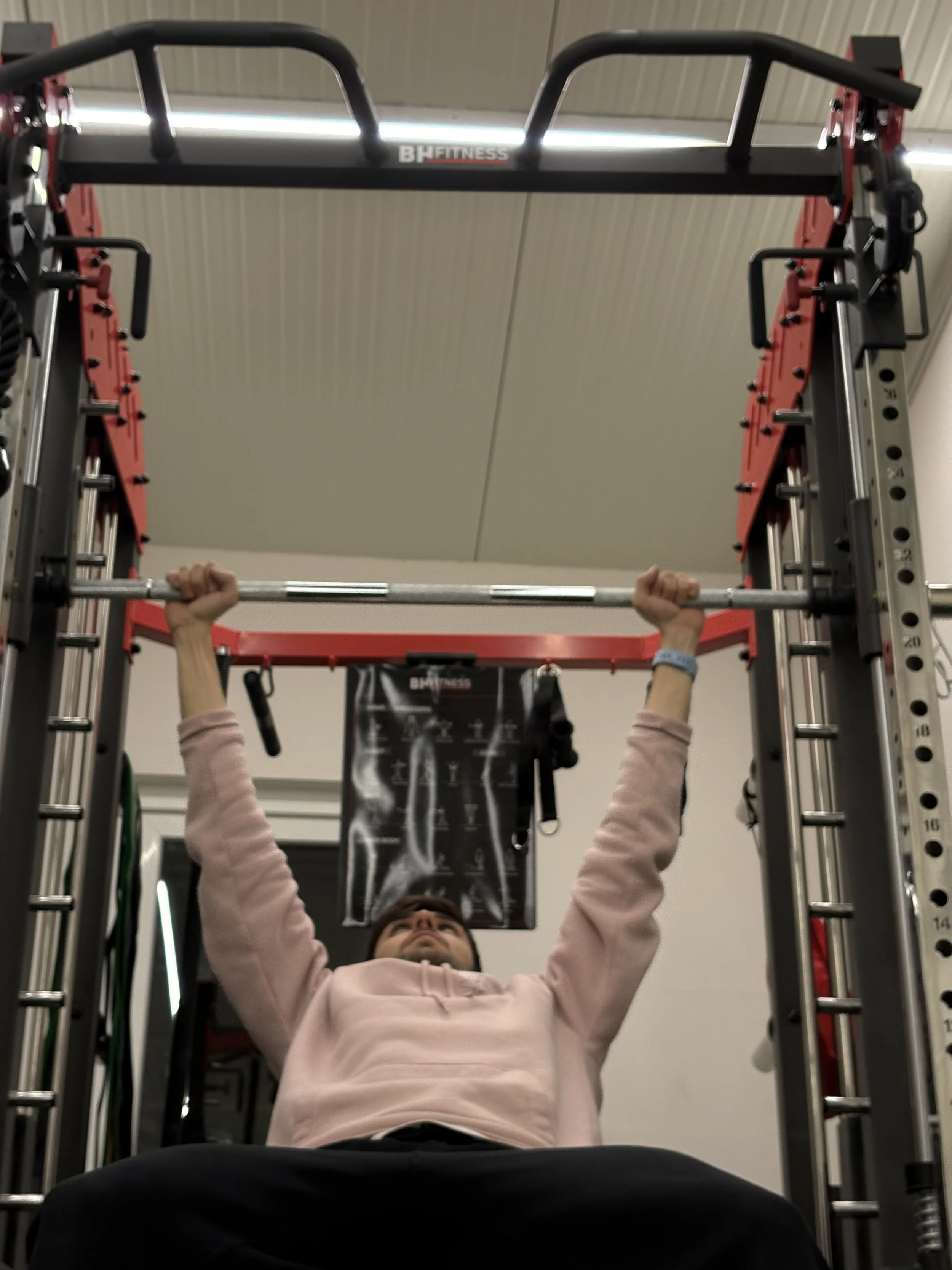 Pessoa deitada no chão de um ginásio, segurando uma barra de peso acima da cabeça, em uma estrutura de academia de musculação.