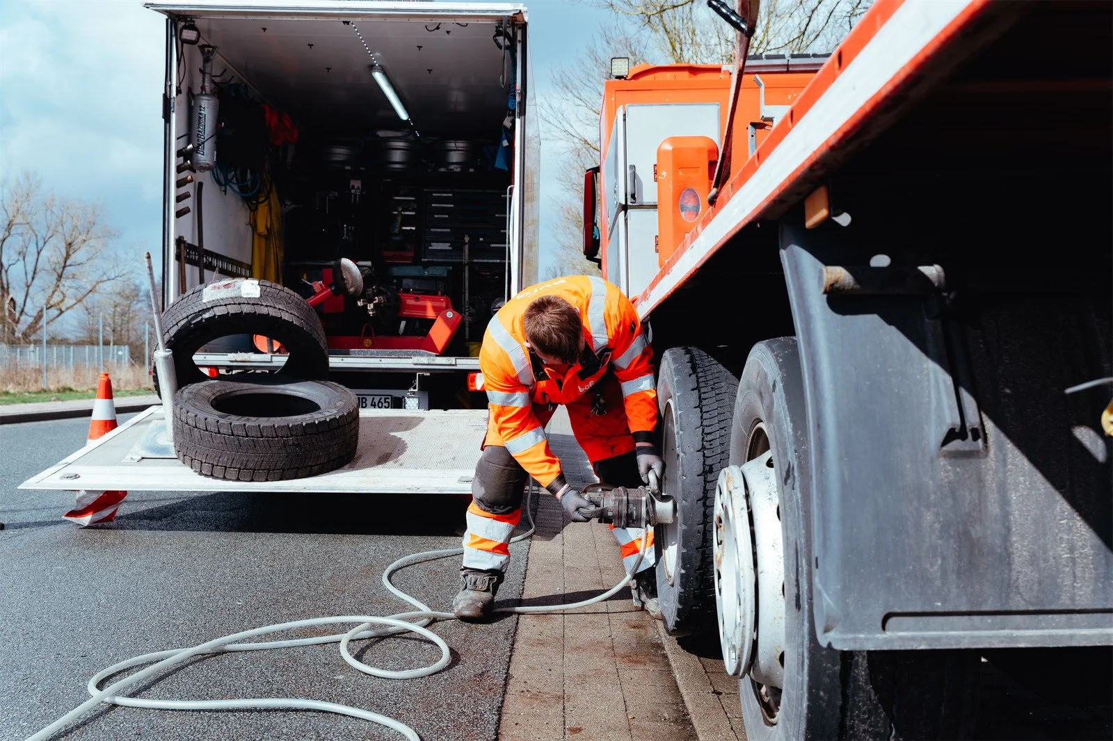Ein Arbeiter in orange Safetykleidung wechselt einen Reifen an einem großen Lkw auf der Straße, mit einem Werkzeug in der Hand und einem Transportwagen im Hintergrund.