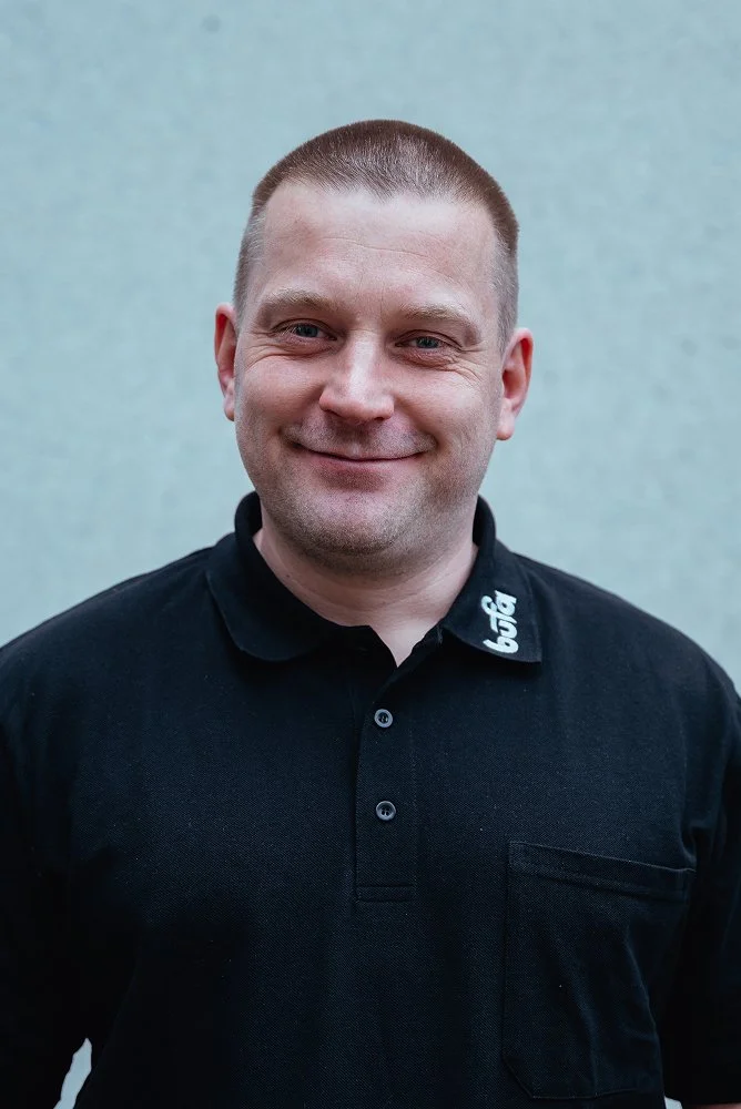 Ein Mann mit kurzen Haarschnitt, trägt ein schwarzes Polo-Shirt mit weißem Logo am Kragen, steht vor einer hellgrauen Wand, lächelt in die Kamera.
