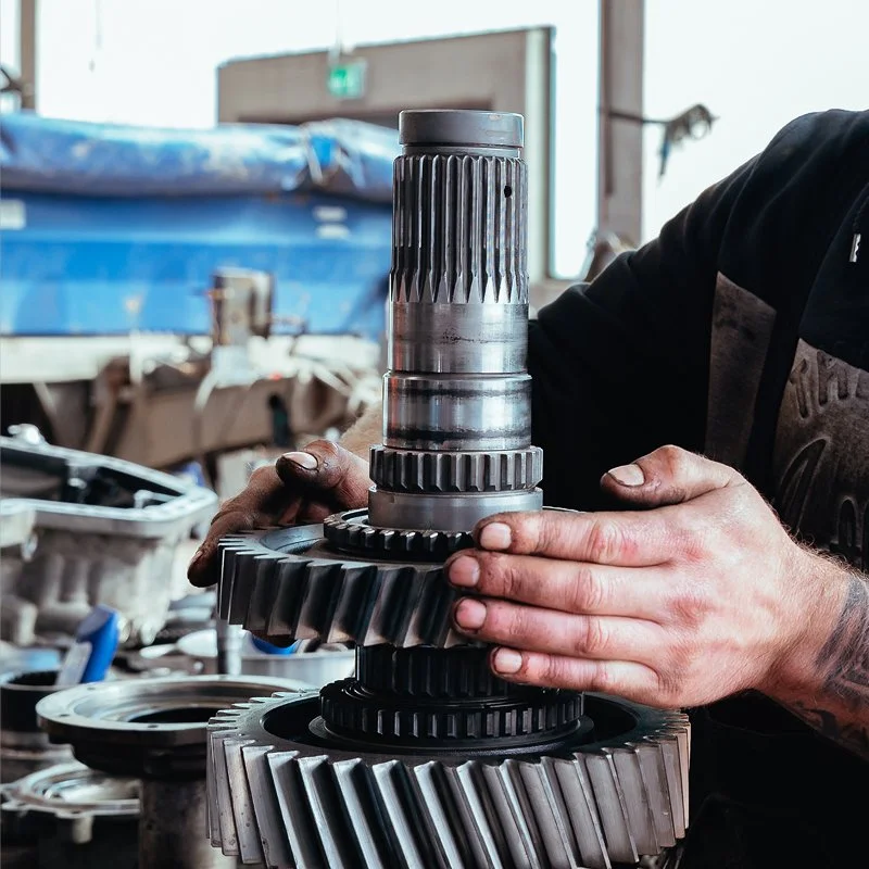 Eine Person inspiziert und hält eine große, metallene Getriebewelle mit Zahnrädern in einer Werkstatt, umgeben von technischen Komponenten.