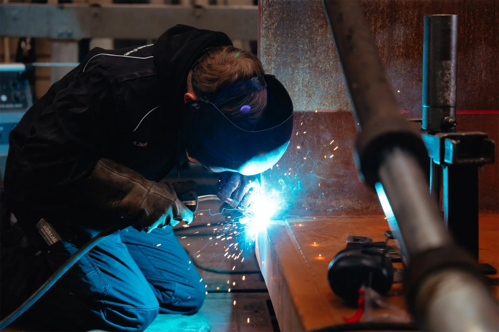 Ein Handwerker schweißt Metall mit Schutzbrille und Schweißhelm, während er auf Knien an einem Werkstück arbeitet, Funken sprühen.
