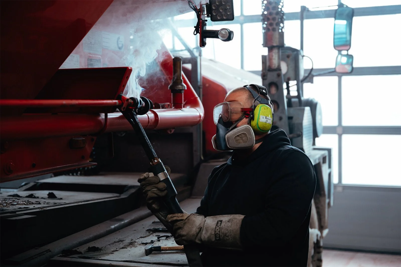 Ein Mann arbeitet mit einem Schweißgerät in einer Werkstatt, trägt Sicherheitsschutzbrille, Gehörschutz und Schutzhandschuhe.