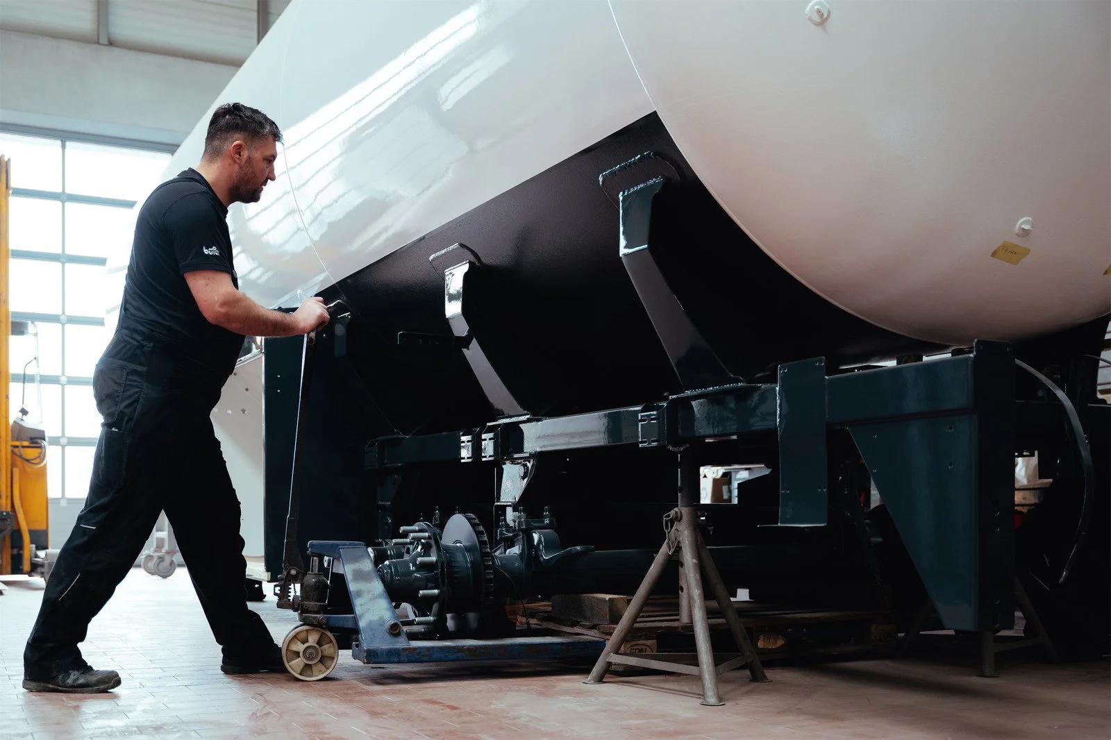 Ein Mann arbeitet an einer großen weißen Boje oder einem Boot im Werkstatt- oder Hangar-Umfeld, das auf einem Gestell steht.