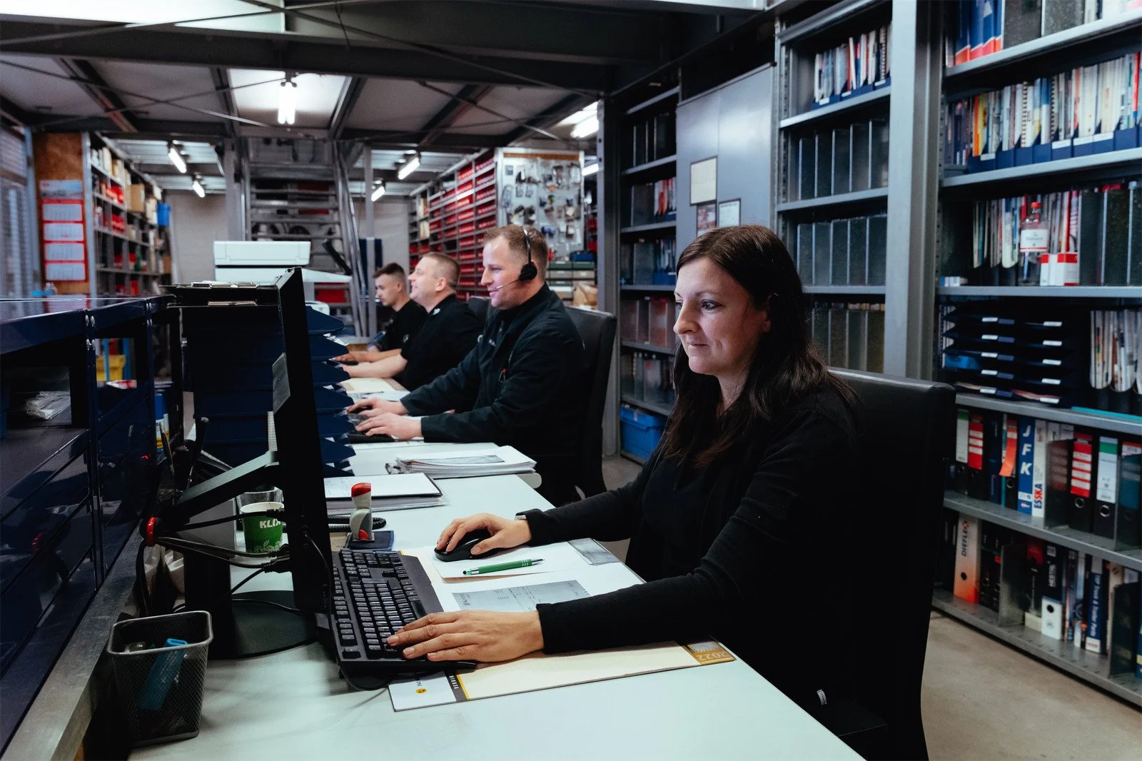Menschen arbeiten in einer Lager- oder Logistikhalle an Computern, gekleidet in schwarze Arbeitskleidung, mit Regalen voller Dokumente und Boxen im Hintergrund.