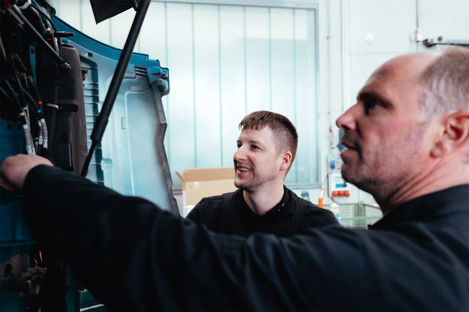 Zwei Männer arbeiten an einem Maschinenraum und betrachten die elektrische Verkabelung, während sie sprechen und lachen.