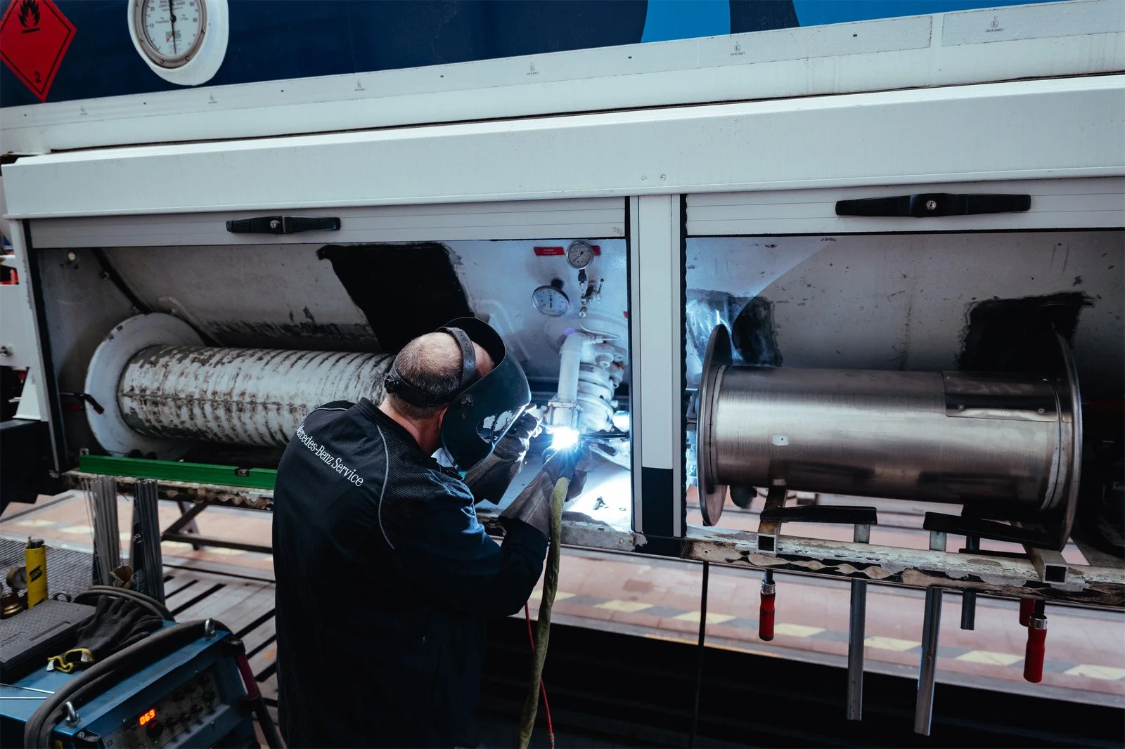 Ein Techniker arbeitet an einem Fahrzeug im Werkstattbereich, trägt Schutzhelm und führt Schweißarbeiten durch, um Teile des Autos zu reparieren.