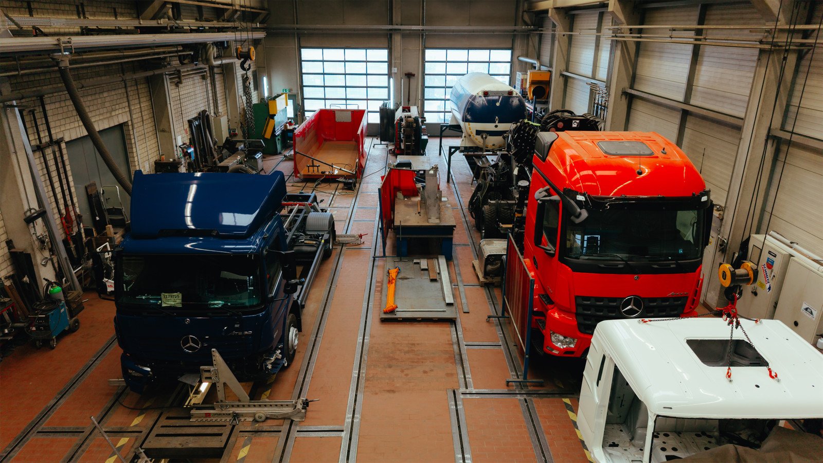 Innenansicht einer Fahrzeugmontagehalle mit mehreren großen LKWs, darunter ein blauer, ein roter und ein weißer LKW, umgeben von Werkzeugen und Ausrüstung.