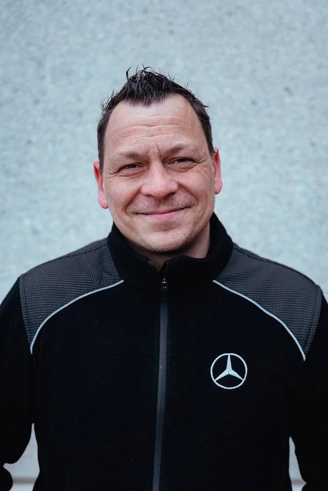 Mann trägt schwarze Jacke mit Mercedes-Benz Logo vor grauer Wand.
