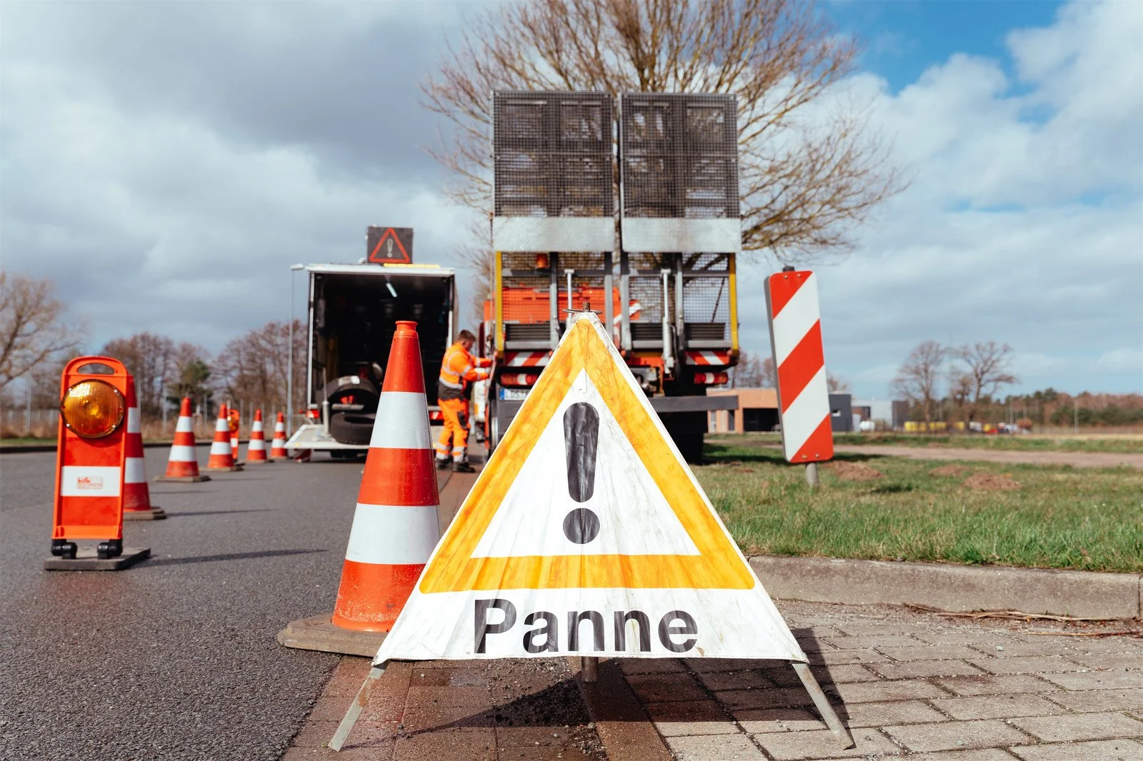 Straßensperrung wegen Panne, Warnschild mit 'Panne', Sicherheitskegel, Arbeiter in orange Warnkleidung, Wartung am Lastwagen, wegen Bauarbeiten absperrbar, sonniges Wetter, Bäume im Hintergrund