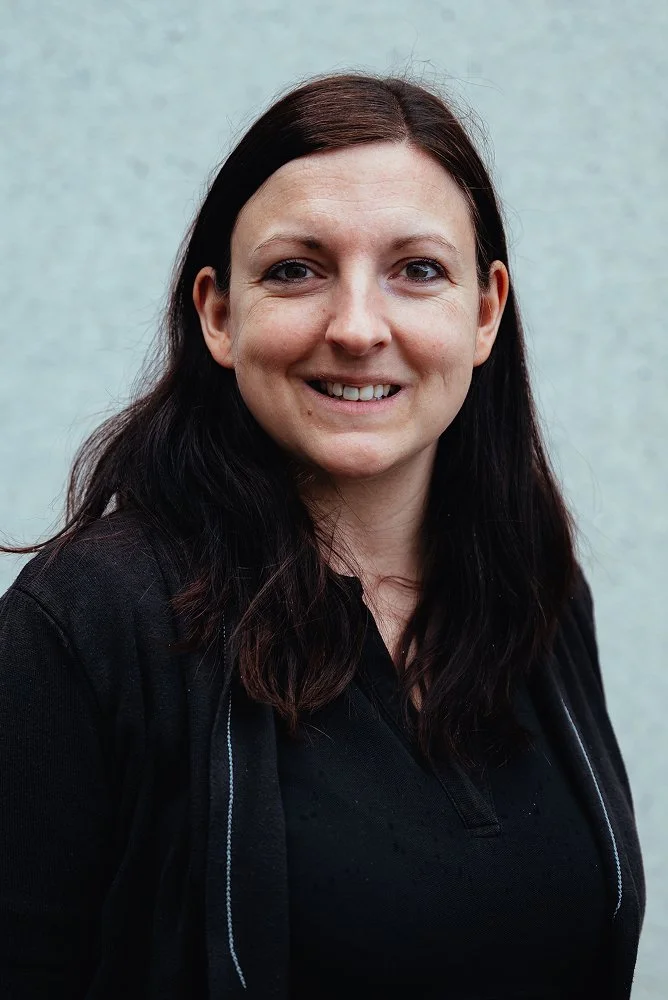 Eine Frau mit braunen, gewellten Haaren lächelnd vor einem hellgrauen Hintergrund, trägt eine schwarze Jacke und ein schwarzes Oberteil.