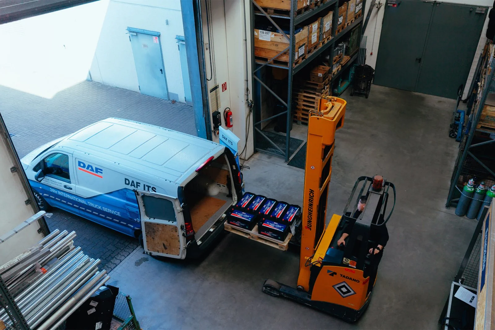 Blick auf eine Lagerhalle, in der eine gelbe Gabelstapler mit einem Mann fährt, der Paletten mit Coca-Cola-Kästen aufnimmt, während ein weißer Lieferwagen mit dem Logo DAF ITS dekoriert ist. Regale mit Kartons und Metallelementen sind sichtbar.