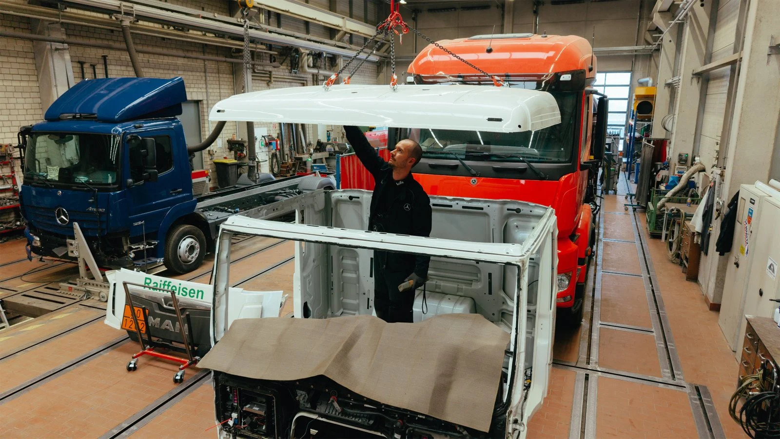 Ein Techniker arbeitet in einer Fabrikhalle mit mehreren Lastwagen und Fahrzeugteilen. Er sitzt in einem weißen Fahrzeugrahmen, während er ein Dachteil eines LKW anbringt.