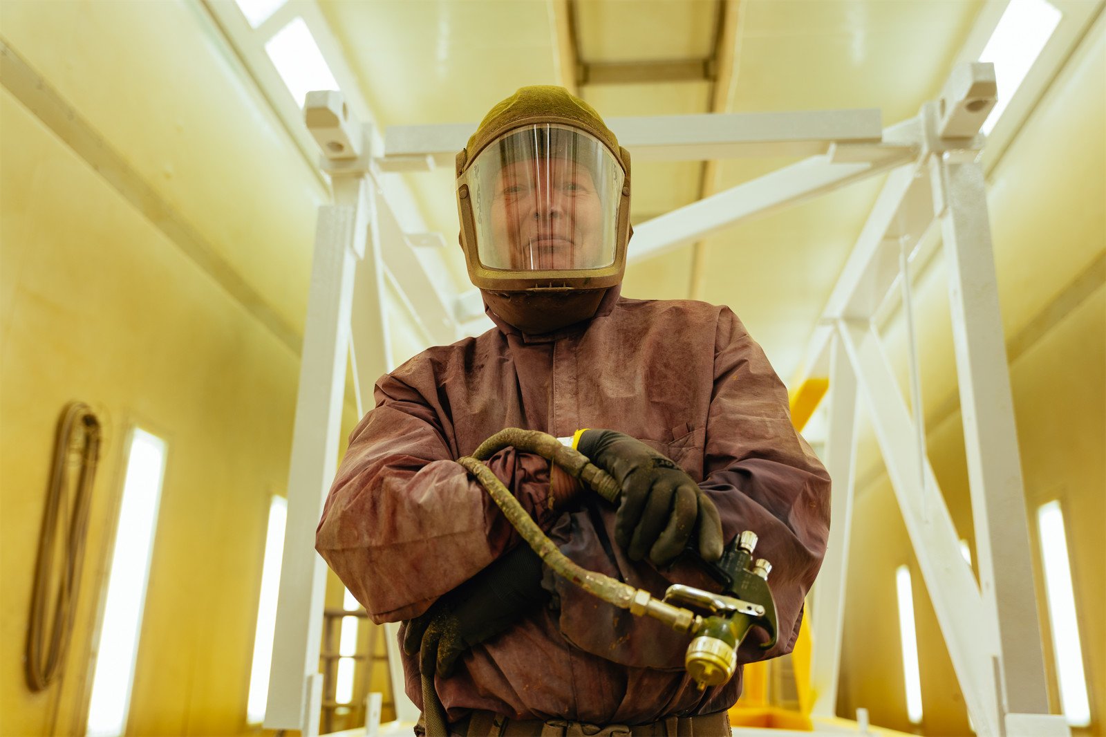 Ein Mann trägt einen Schutzhelm und arbeitet mit einem Druckluftschlauch in einem industriellen Umfeld, vermutlich in einer Fertigungshalle.