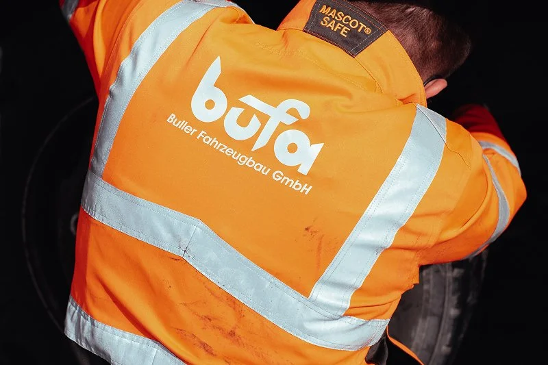 Blick auf eine Person in einem orangefarbenen Sicherheitsjacke mit reflektierenden Streifen, die auf einen Reifen schaut. Das Jacke hat das Logo 'bufa' und den Text 'Buller Fahrzeugbau GmbH'.