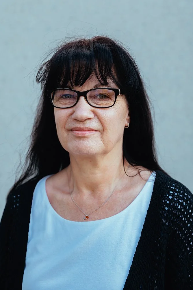 Porträt einer Frau mit schwarzen Haaren, Brille und dezentem Lächeln, trägt ein weißes T-Shirt und eine schwarze Strickjacke vor einem grauen Hintergrund.