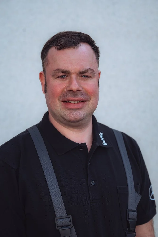 Ein Mann trägt ein schwarzes Polo-Shirt mit Logos und trägt schwarze Schultergurte, steht vor einer hellen Wand.