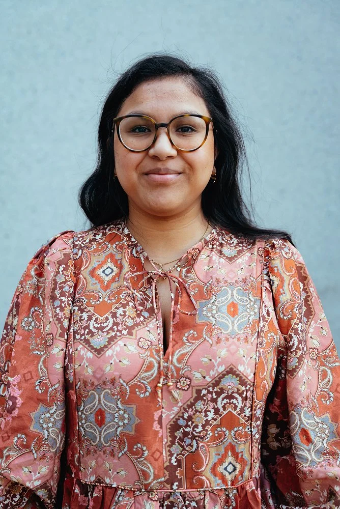 Eine Frau mit dunklen Haaren, Brille und einem floral gemusterten Kleid auf grauem Hintergrund.