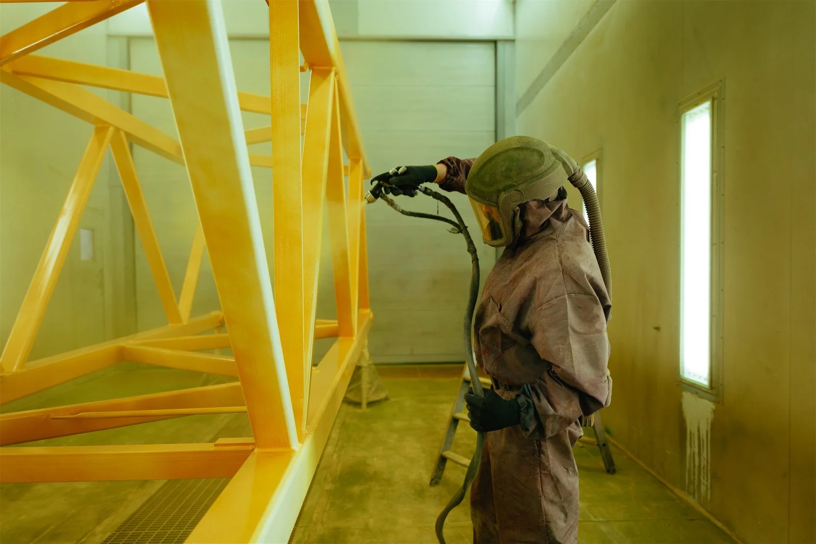 Ein Arbeiter in Schutzkleidung und Helm sprüht ein großes, gelbes Metallgerüst in einer industriellen Umgebung an.