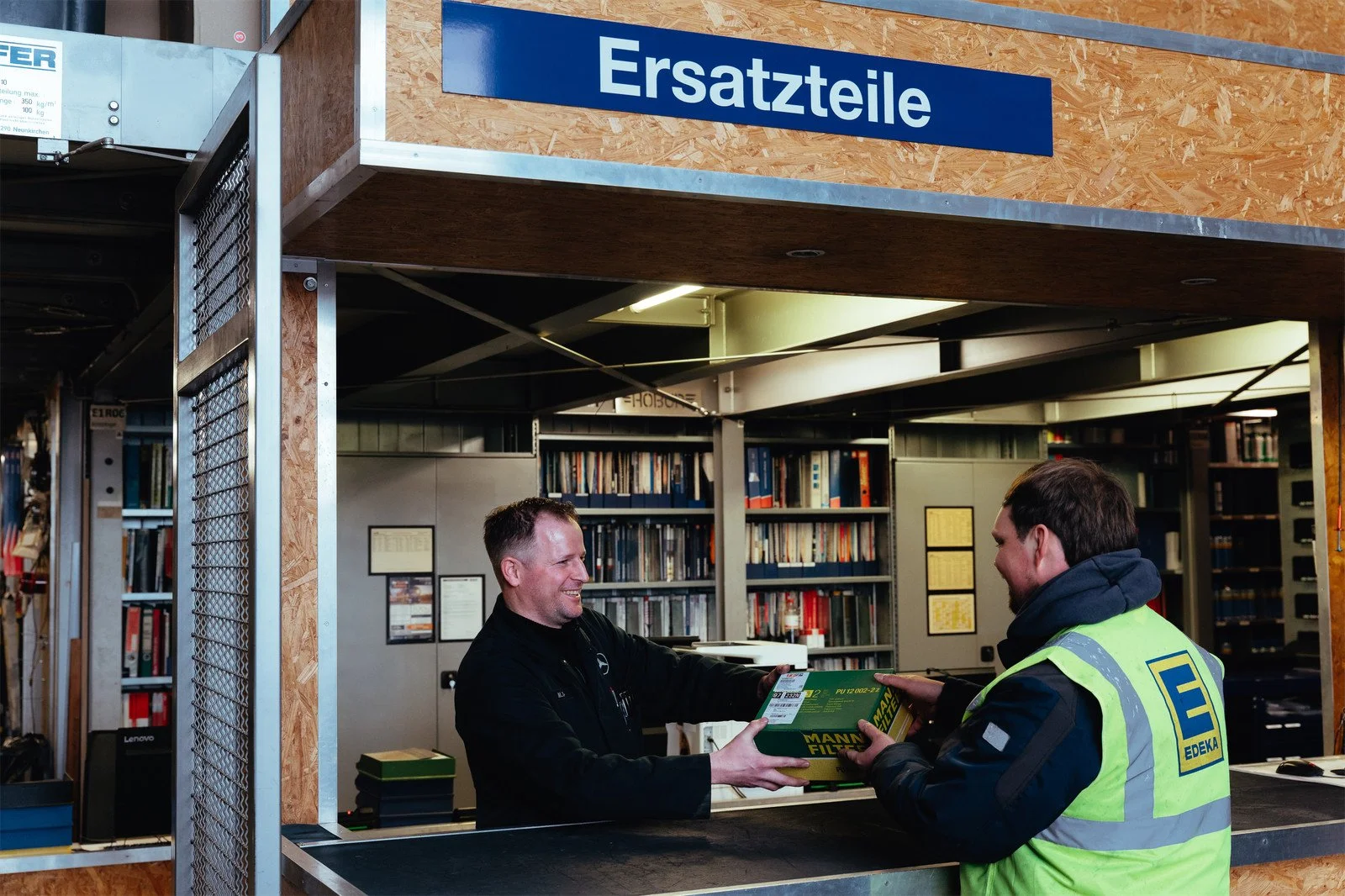 Zwei Männer in einem Lager mit Regalen voller Bücher und Akten, der eine Mann trägt eine grüne EDEKA-Reflektorweste, der andere hält eine Packung Reifenfilter, beide lächeln und tauschen die Ware aus.