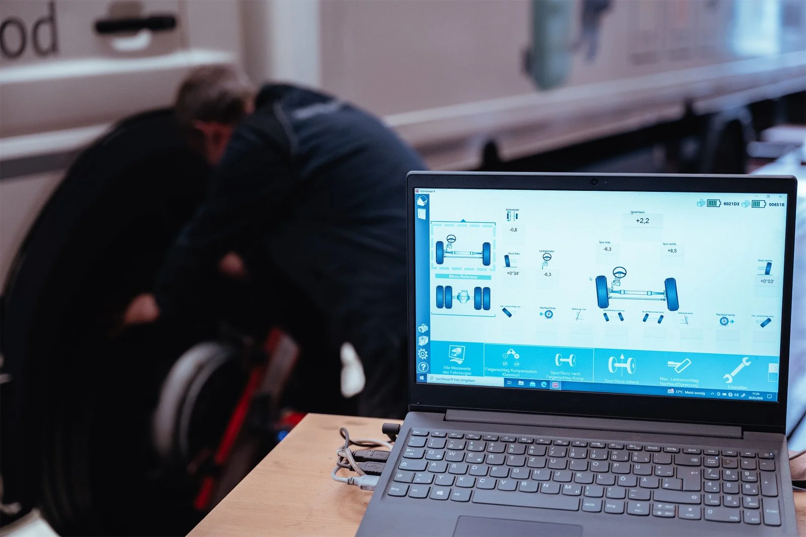 Laptop zeigt eine Fahrzeugdiagnose mit Achs- und Federdämpfer-Einstellungen, im Hintergrund sitzen zwei Männer und diskutieren.
