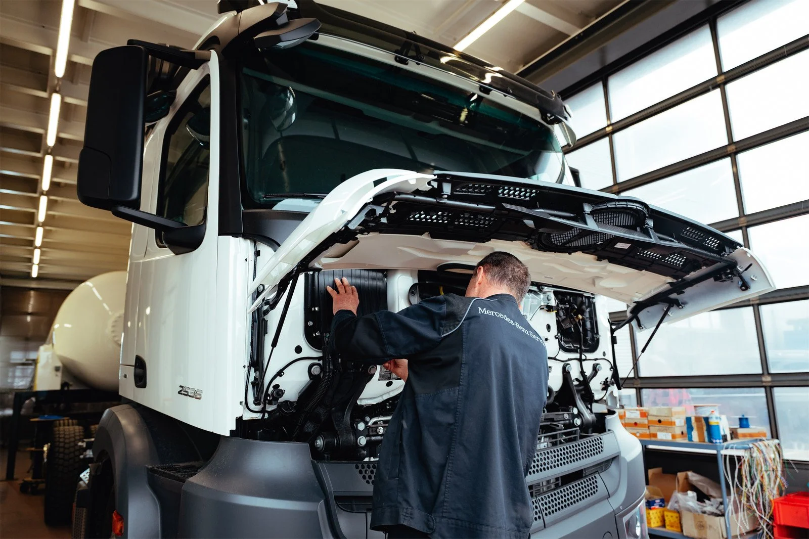 Ein Techniker arbeitet in einer Werkstatt an einem weißen Mercedes-Benz LKW mit ausgehobenem Motorhaube, vermutlich bei einer Wartung oder Reparatur.