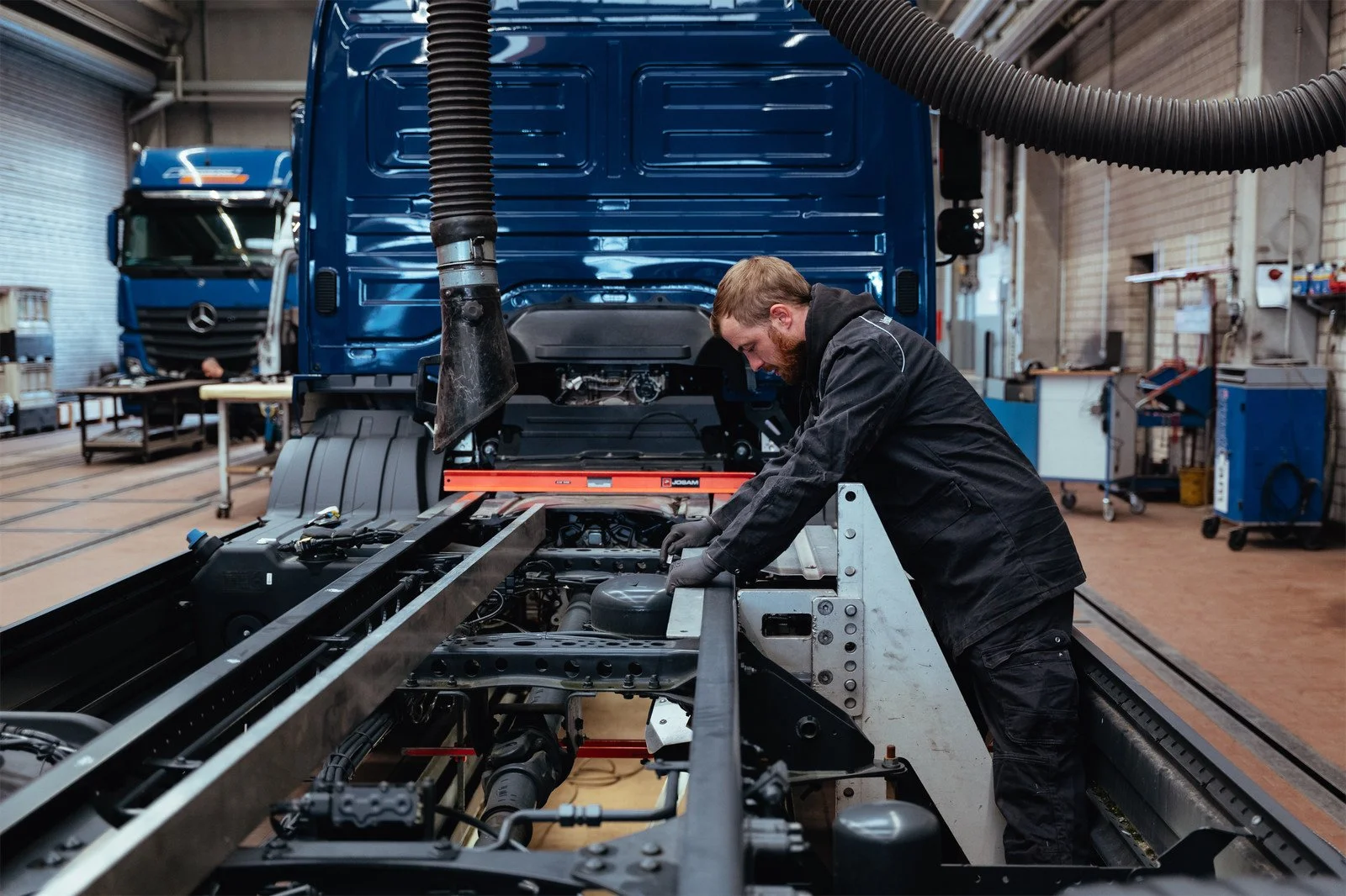 Ein Mann arbeitet in einer Werkstatt an einem großen LKW, der auf einem Reparaturständer steht, mit verschiedenen Werkzeugen und Ausrüstung im Hintergrund.