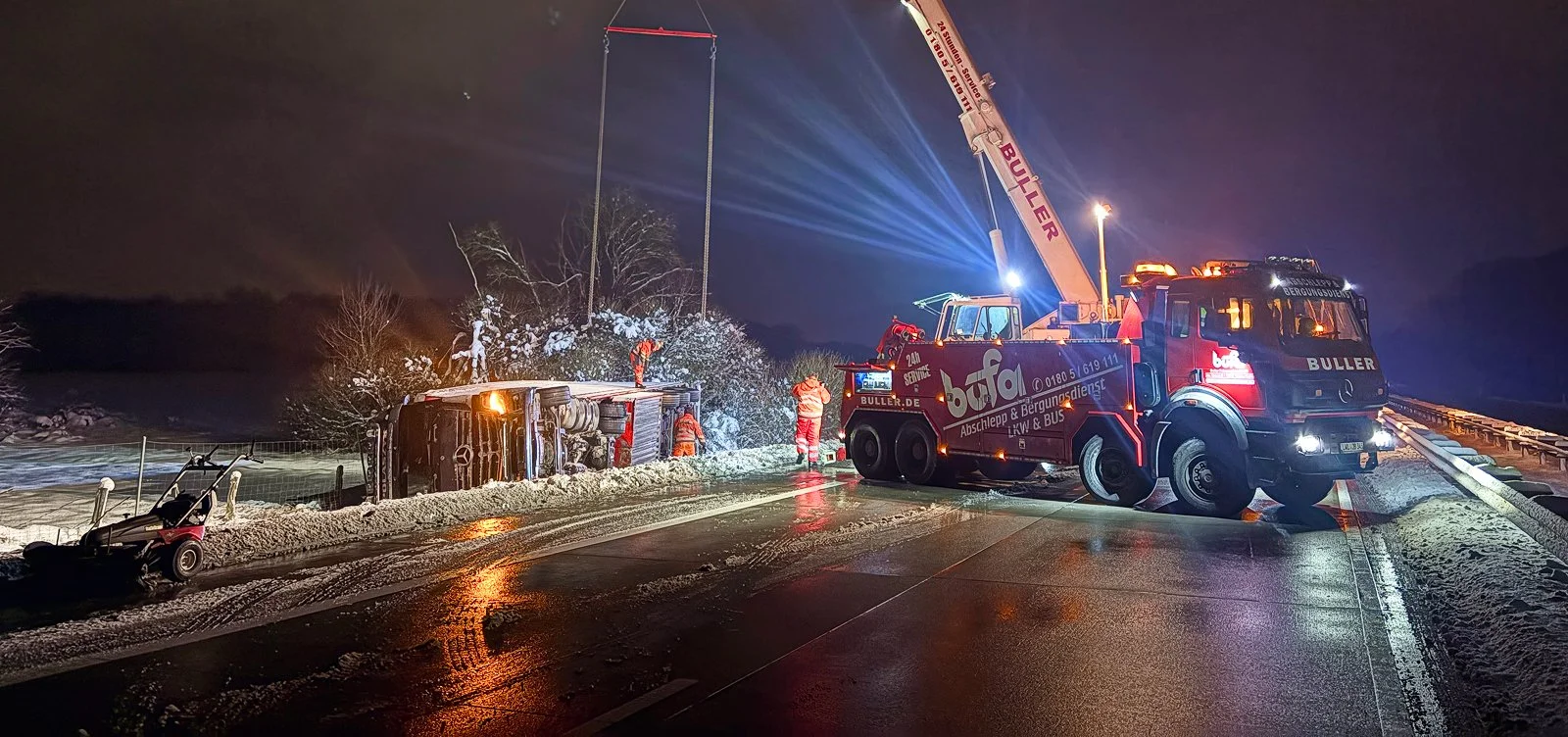 Ein Unfall auf einer verschneiten Straße bei Nacht, bei dem ein umgestürztes Fahrzeug zu sehen ist, das von einem Abschleppwagen geborgen wird, während Einsatzkräfte im Einsatz sind.