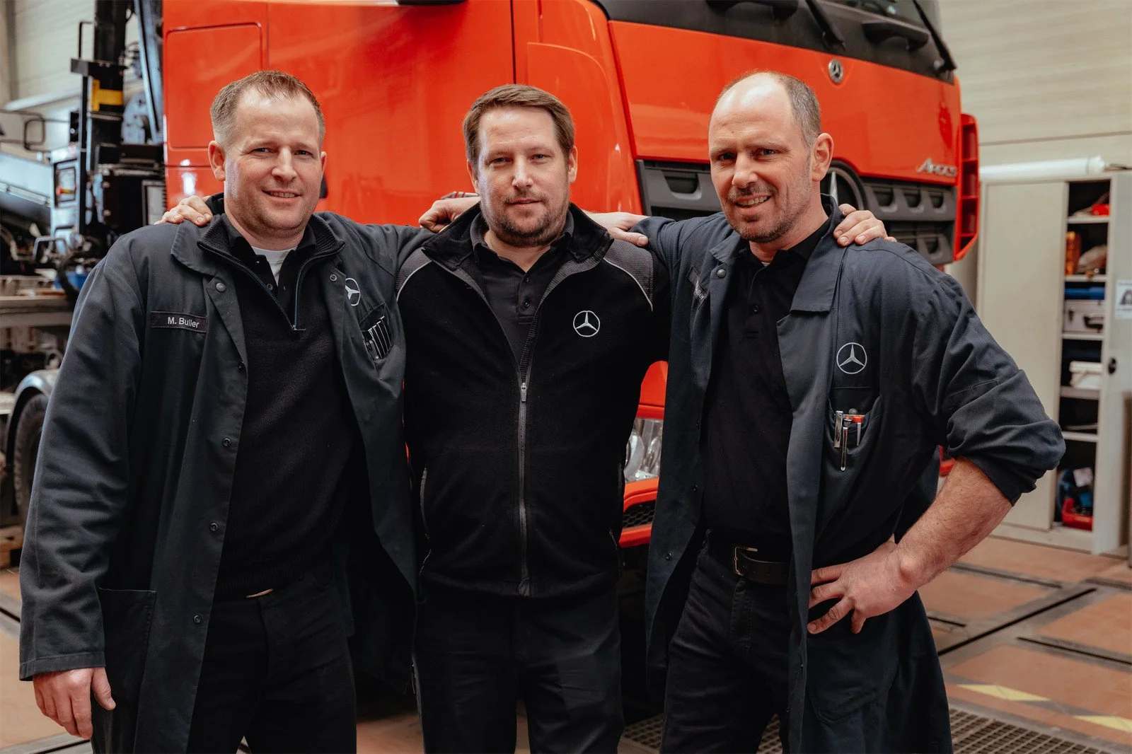 Drei Männer in Werkstatt stehen vor einem orangefarbenen Lkw, tragen dunkle Arbeitskleidung mit Mercedes-Benz-Logo, lächeln und haben die Arme umeinander geschlungen.