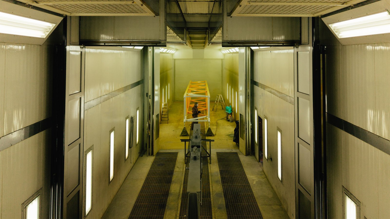 Innenansicht einer großen, weißen, industrialartigen Halle mit einem gelben Rahmen, der auf einer Hebebühne oder einem Kran montiert ist, und einigen Arbeitern, die daran arbeiten.