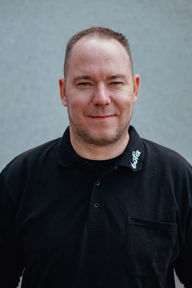 Ein Mann mit kurzen Haaren und Bart, trägt ein schwarzes Polo-Shirt, steht vor einer grauen Wand.