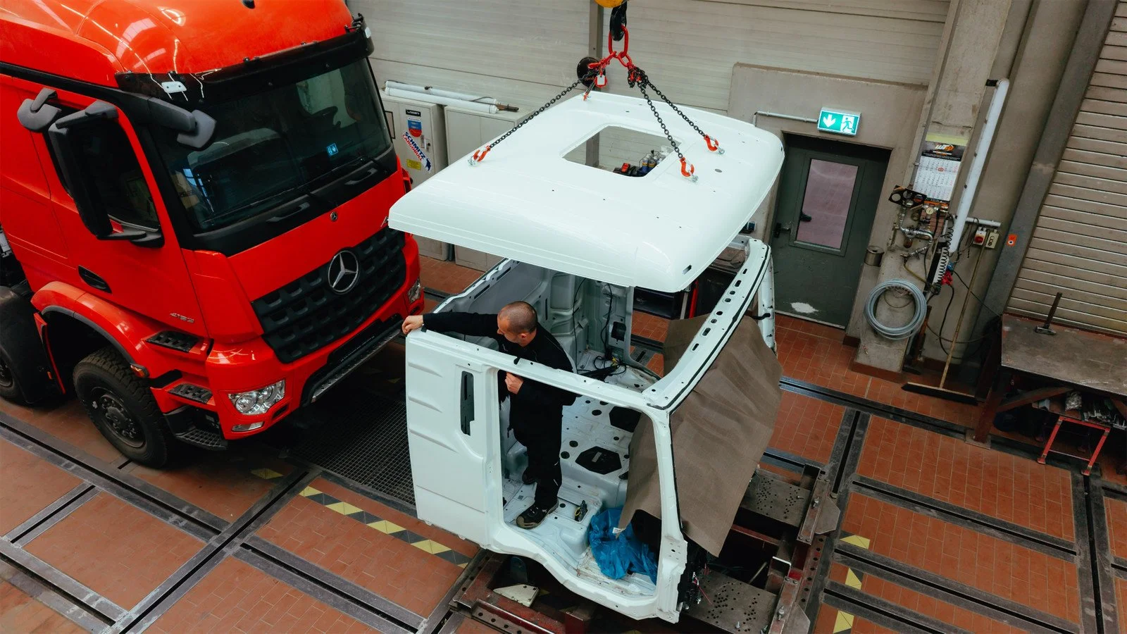 Ein Monteur arbeitet an der Karosserie eines weißen Fahrzeugs in einer Werkstatt, während ein roter LKW neben ihm steht.