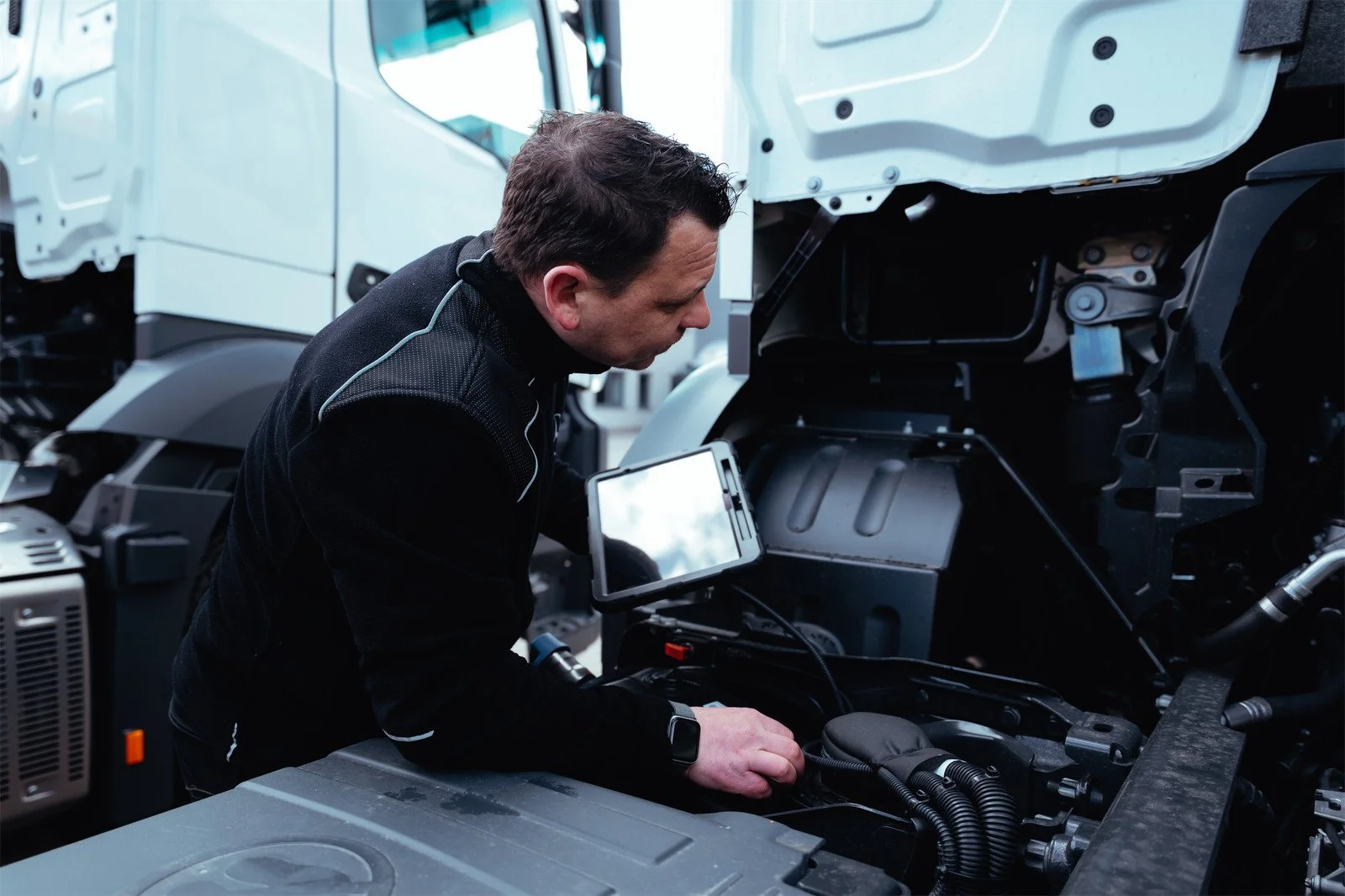 Mann arbeitet an einem Fahrzeugmotor, während er eine Diagnose oder Reparatur durchführt, mit einem Reparatur-Tablet in der Hand.