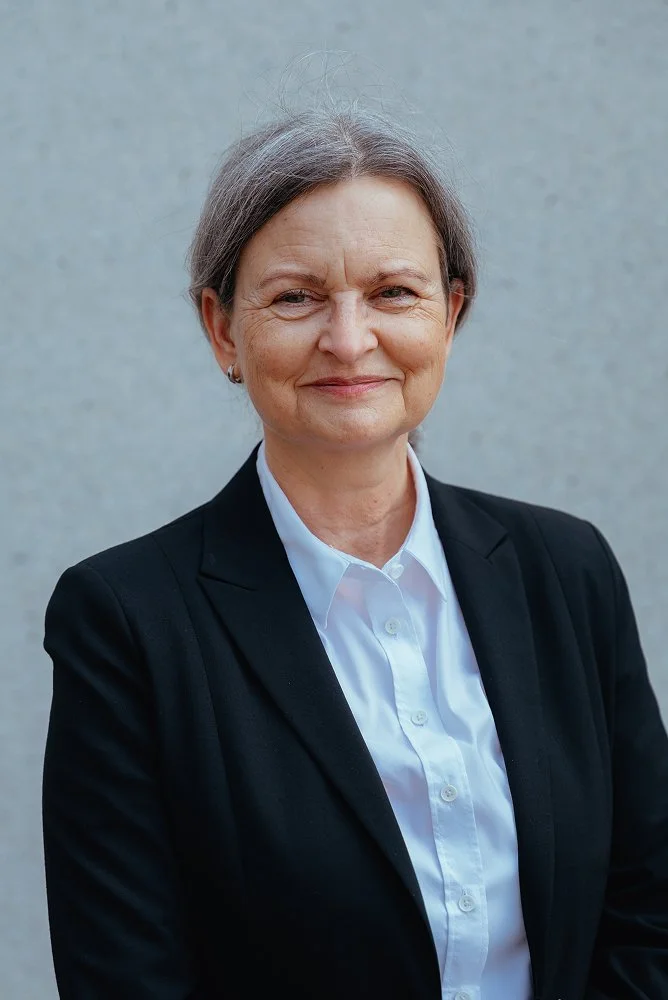 Porträt einer Frau in formeller Kleidung vor grauem Hintergrund, lächelnd.
