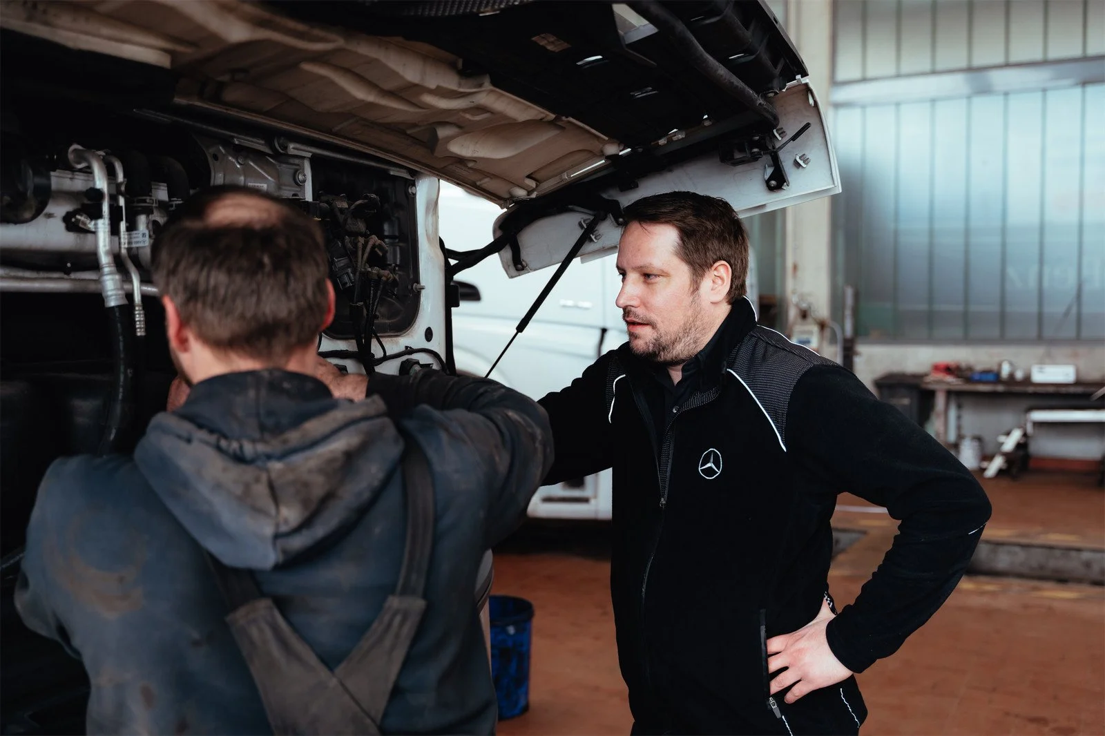 Zwei Männer in einer Werkstatt reparieren ein Fahrzeug, einer zeigt auf den Motor, der andere hört zu.
